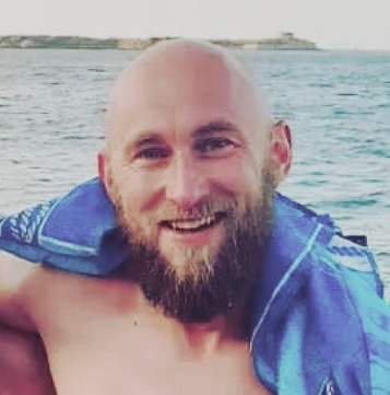 Smiling man with a beard, draped in a blue towel, standing by the sea.