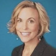 Portrait of a smiling person with short, blonde hair, wearing a suit jacket, in front of a blue background.