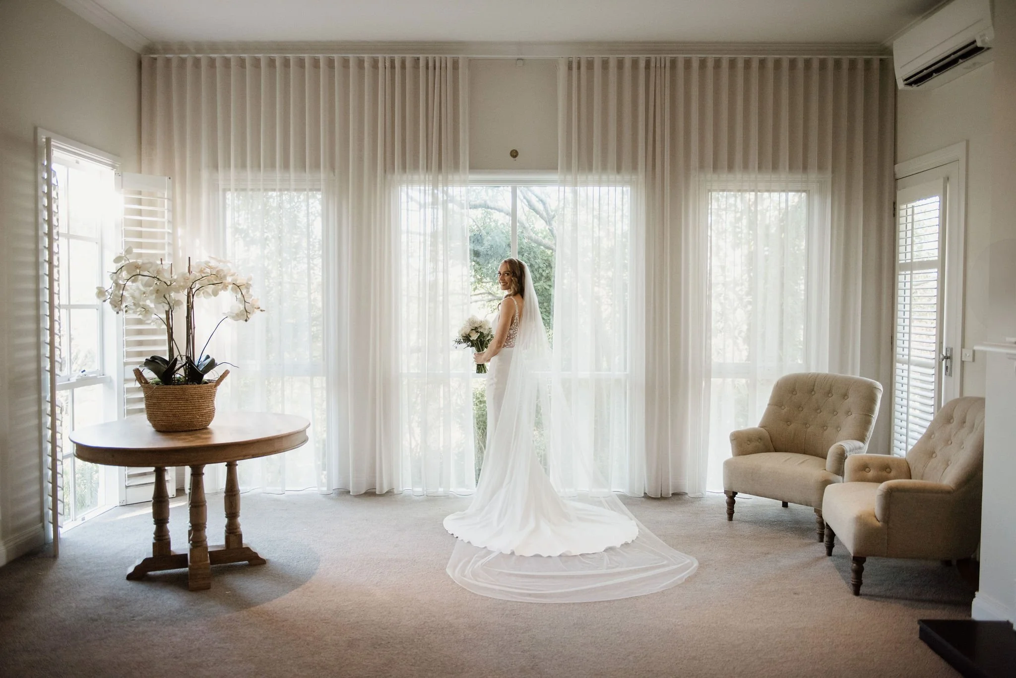A woman in a wedding dress holding a bouquet, standing in front of large windows with sheer curtains, in a well-lit living room with beige chairs, a round wooden table with white orchids, and an air conditioning unit.
