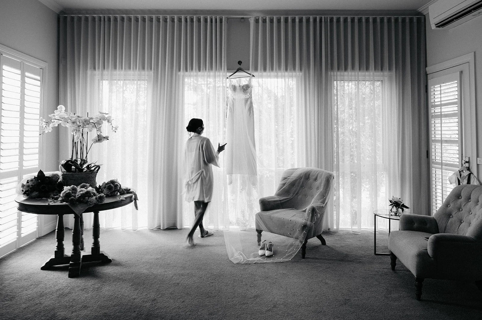 A woman looks at a wedding dress hanging in front of a large window in a bright room. The room has furniture, flowers on a table, and sunlight filtering through curtains.