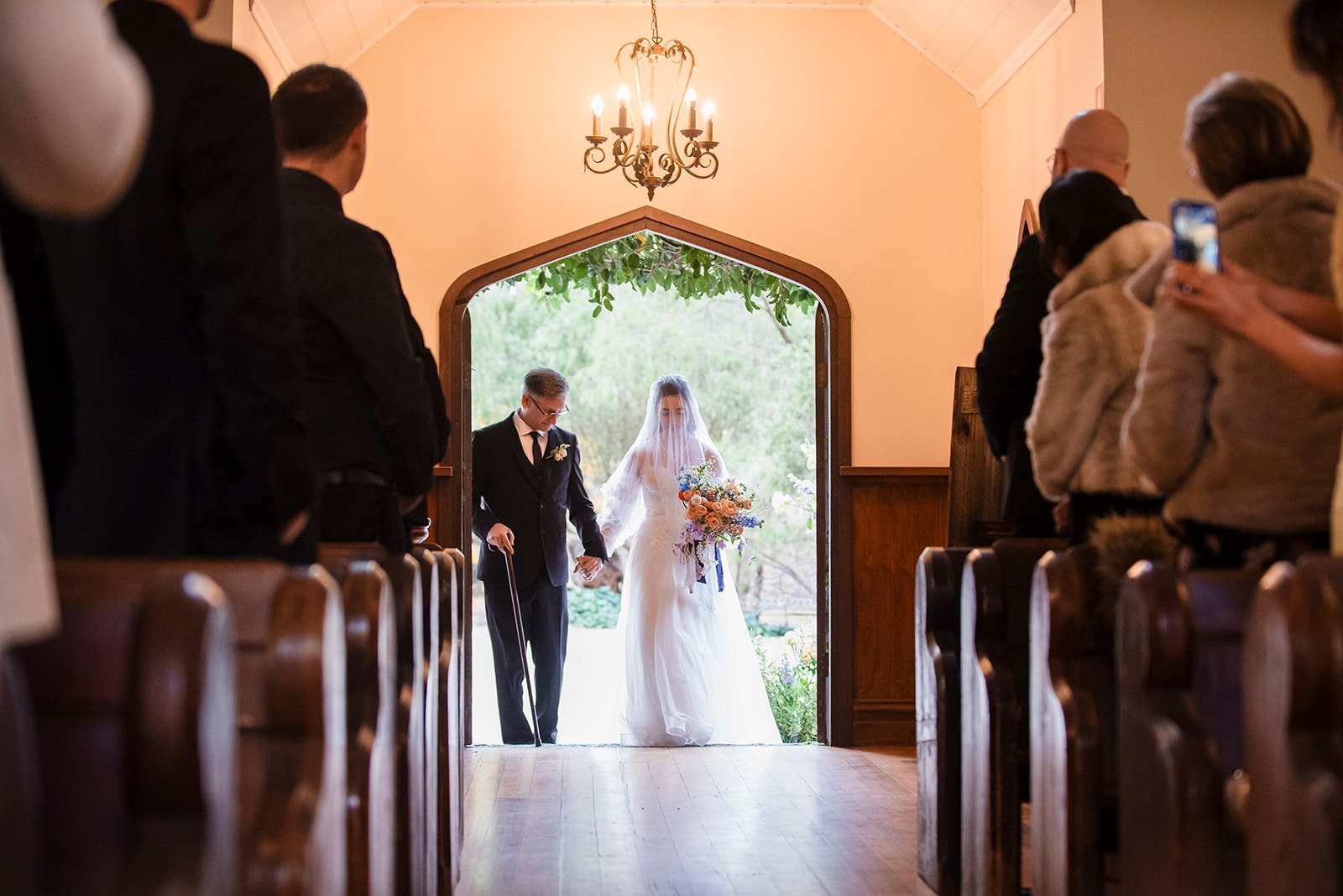 A bride and groom walking out of a church, holding hands, with guests on either side of the aisle watching and taking pictures. The bride is wearing a white wedding dress with a veil and holding a bouquet, and the groom is in a black suit with a cane.