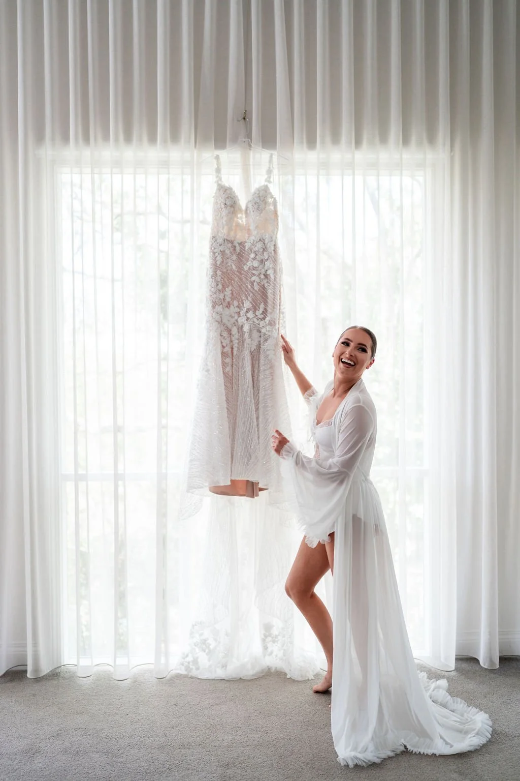 A smiling woman in a white robe standing next to a wedding dress hanging in front of sheer curtains in a bright room.