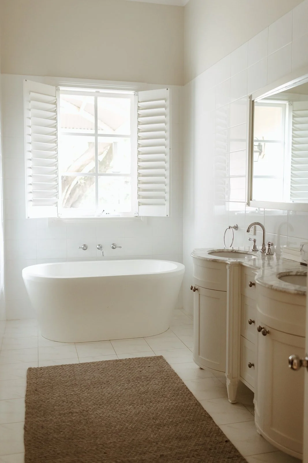 Bright bathroom with a bathtub under a window with white shutters, a double vanity with marble countertop and two sinks, and a mirror above.