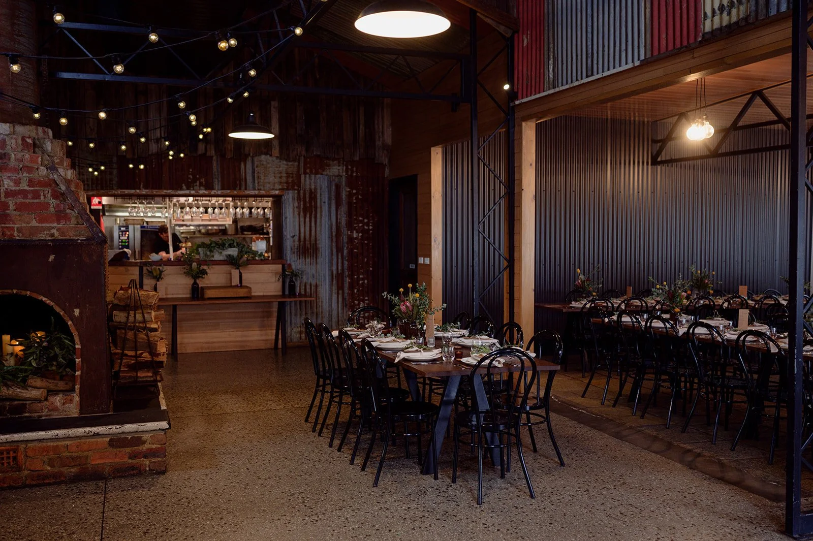 Interior of a rustic restaurant with wooden and brick walls, decorated with string lights. There are multiple tables set for dining with floral centerpieces, surrounded by black chairs. The space features warm lighting and a visible bar area in the background.