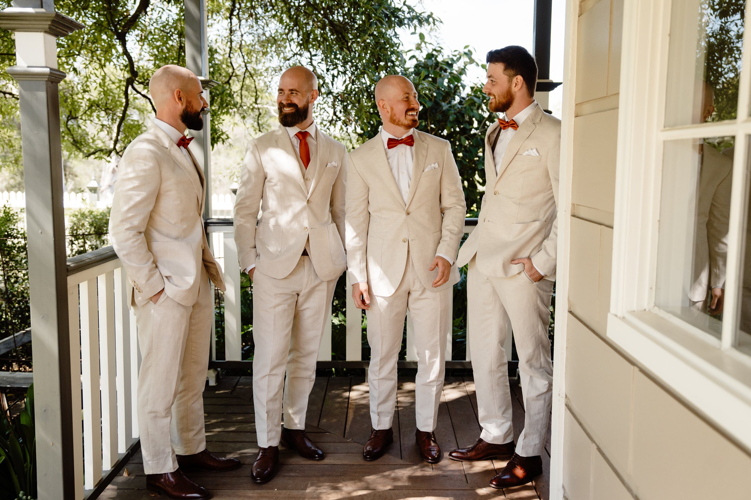 Five men dressed in beige suits with red bow ties standing on a porch, smiling and talking, with greenery in the background.