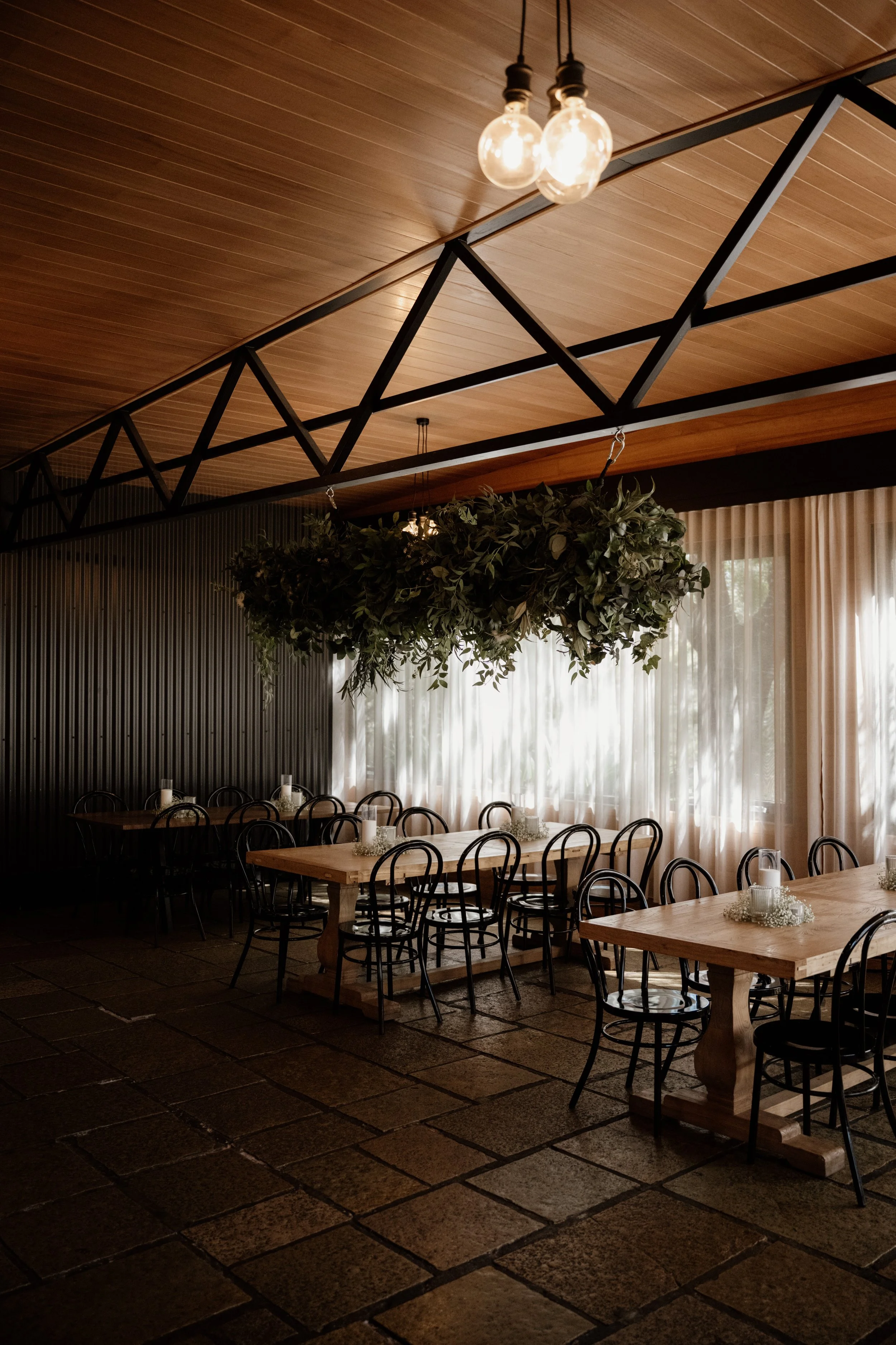 A dining area with wooden tables and black chairs, decorated with candles and flowers, featuring hanging light bulbs and greenery, and large windows with curtains letting in natural light.