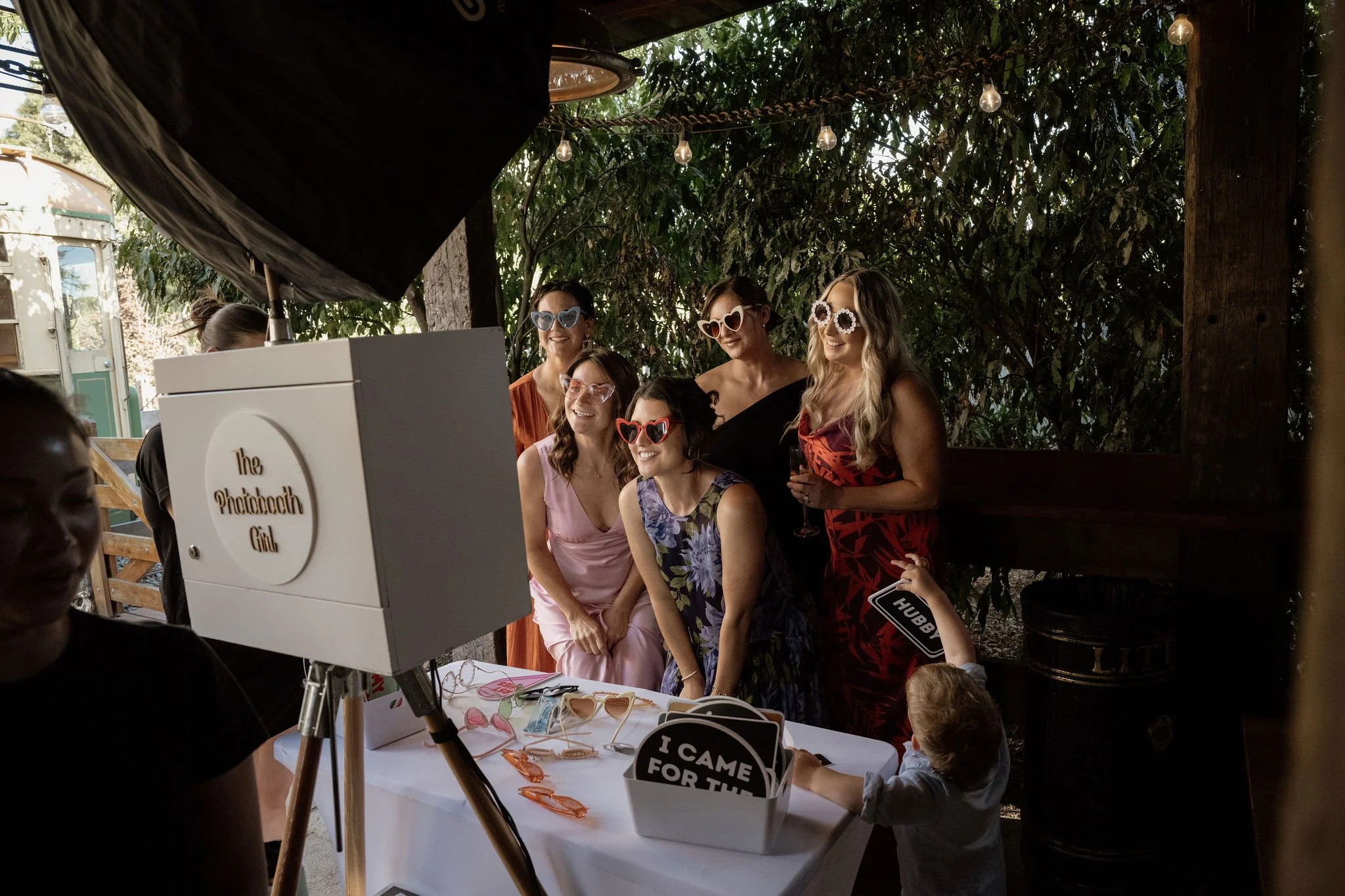 Group of women wearing colorful sunglasses, posing for a photo at a booth labeled 'The Photobooth Girl' at an outdoor event, with green trees and string lights in the background.