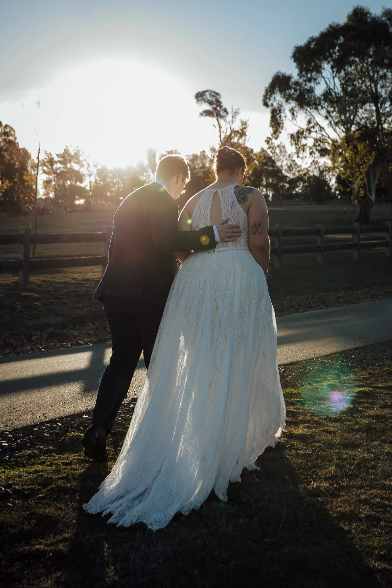 A couple walking outdoors, with the sun setting behind them, dressed in wedding attire, the bride in a white lace gown and the groom in a suit.