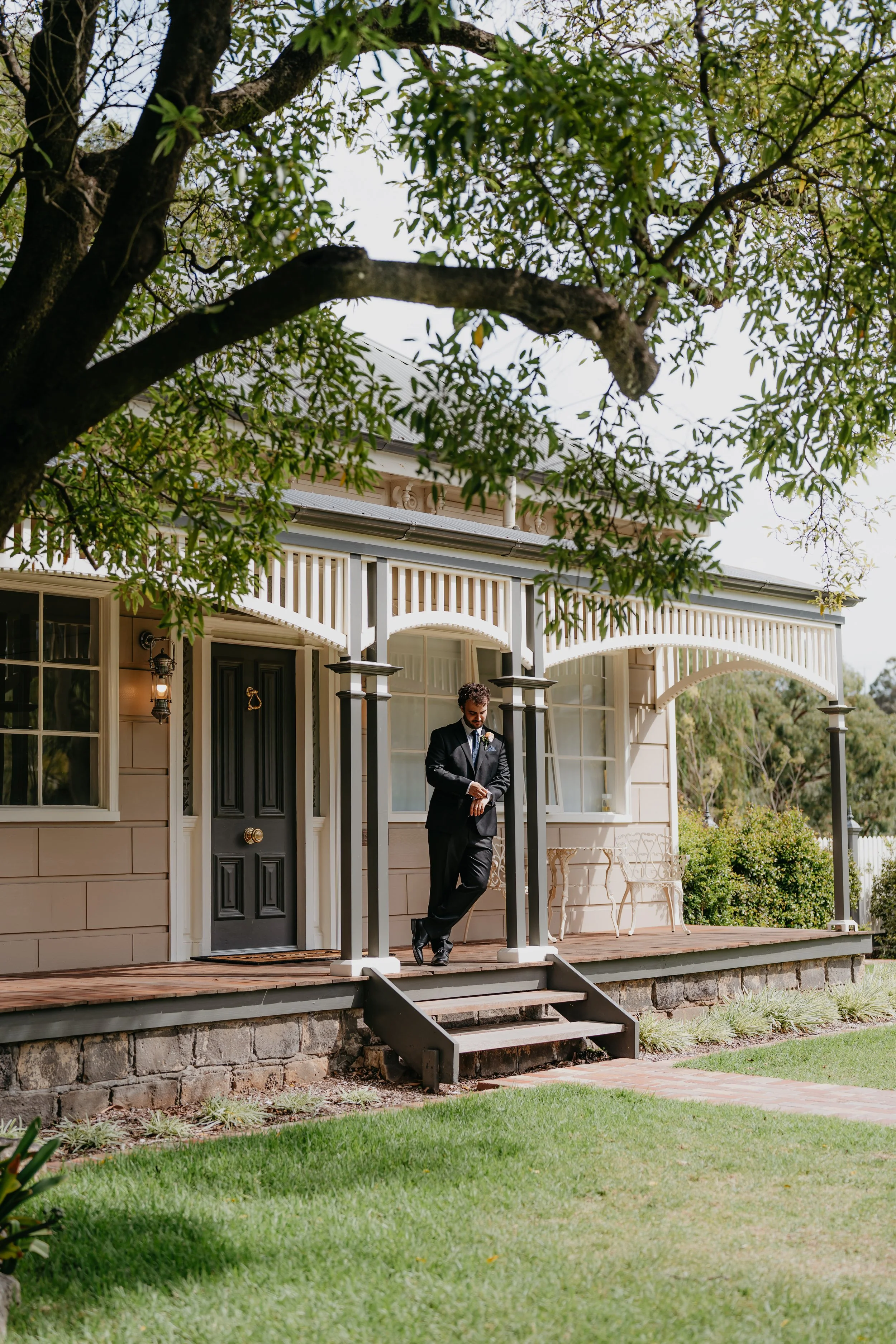 Man in a suit standing on porch of a house with a front door, stairs, and white railings, surrounded by trees and green lawn.