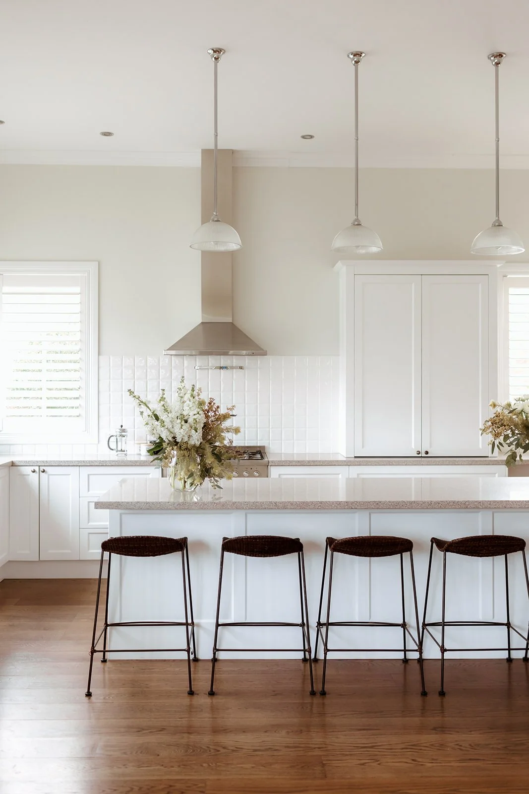 Modern white kitchen with a marble island and four black barstools, flowers on the counter, and pendant lights hanging from the ceiling.