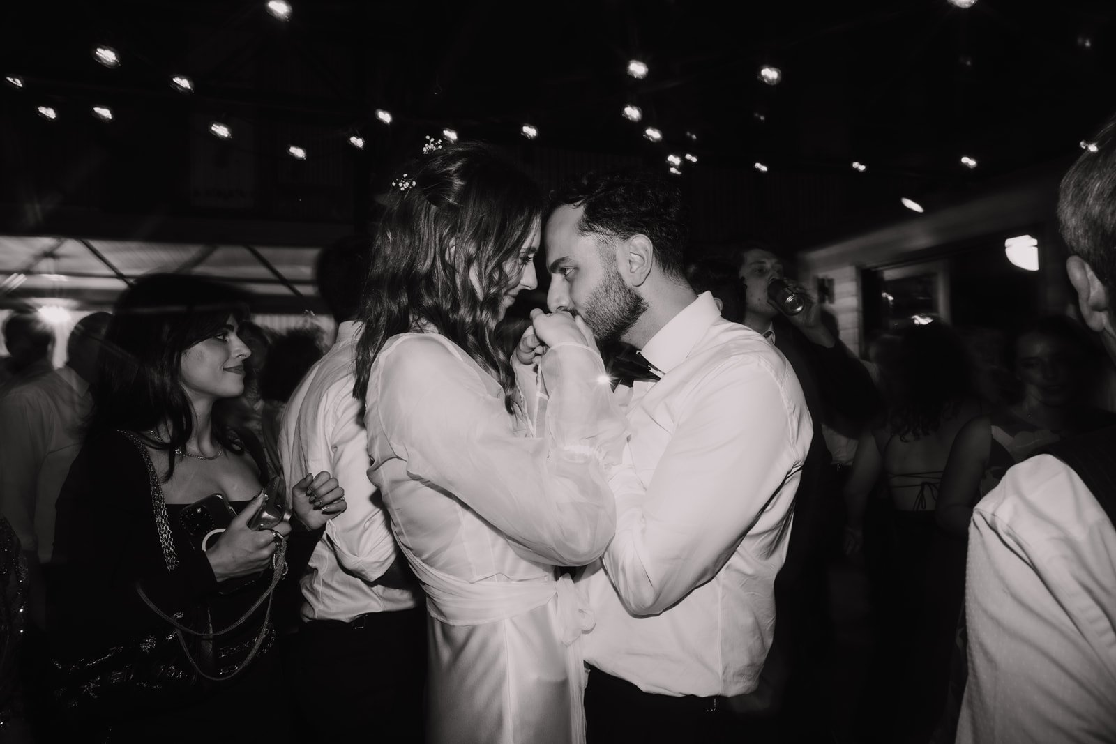 Black and white photo of a man and woman dancing closely, their foreheads touching, during a celebration or wedding reception. They are surrounded by other guests, some holding drinks, in a dimly lit indoor setting.