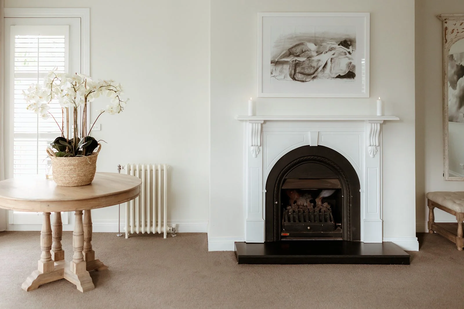 Living room with a white fireplace, black fire grate, and a large framed black-and-white artwork above. There are two white candles on the fireplace mantel. To the left is a round wooden table with a potted orchid in a woven basket. Light flows in from a window slatted door on the left, and beige carpet covers the floor.