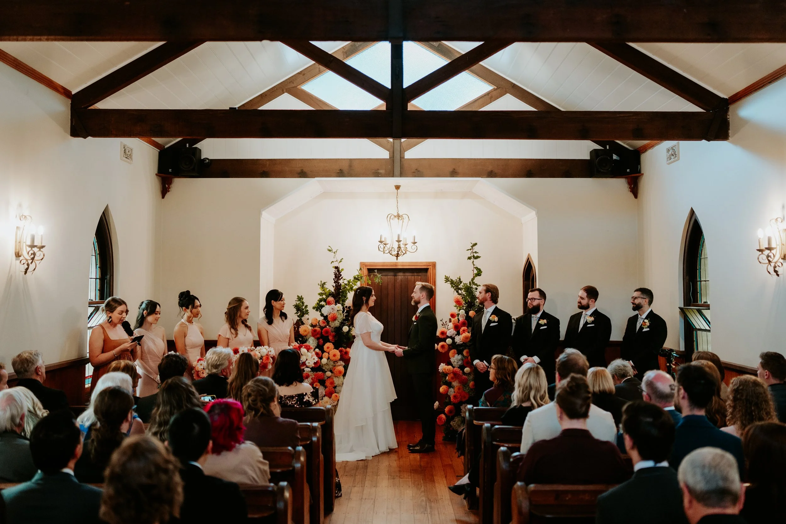 A wedding ceremony taking place inside a church with guests seated facing the altar. The bride and groom are holding hands in front of a floral arrangement, with bridesmaids and groomsmen standing on either side. The church has high ceilings with wooden beams and arched windows.