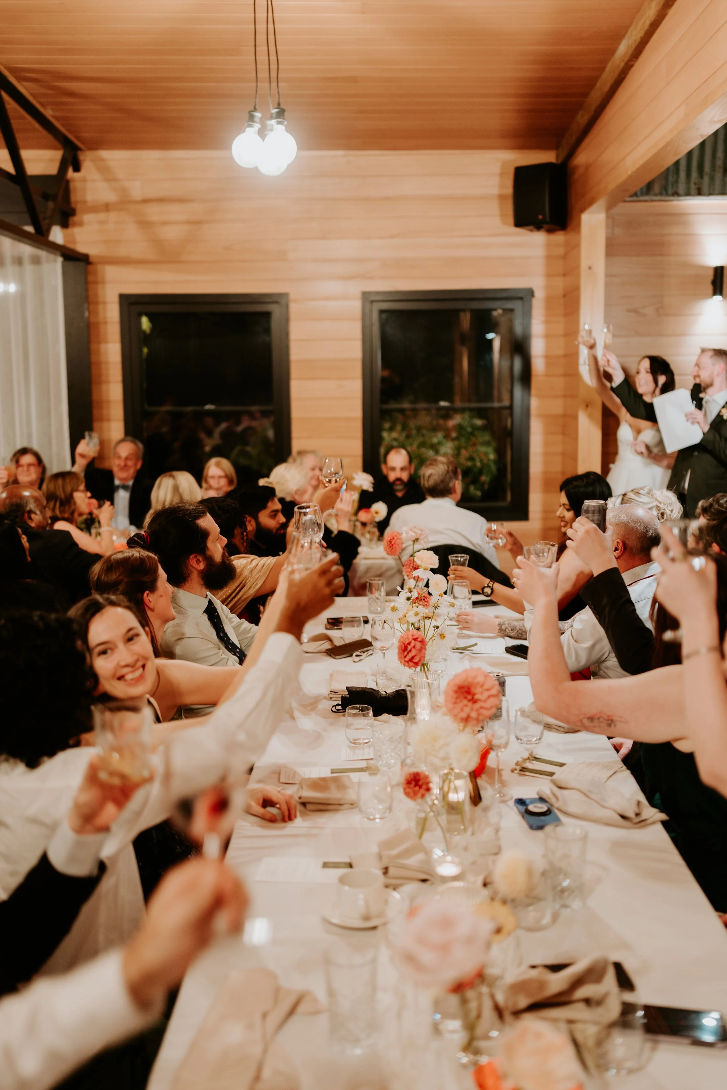 A large group of people attending a wedding reception in a warmly lit wooden hall, raising glasses in a toast, with a bride and groom standing on a small stage.