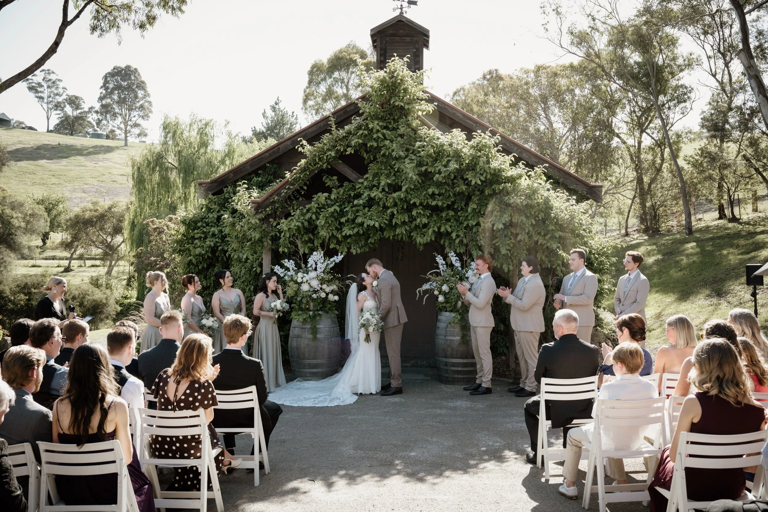 A wedding ceremony outdoors with the bride and groom kissing under a decorated archway surrounded by guests.