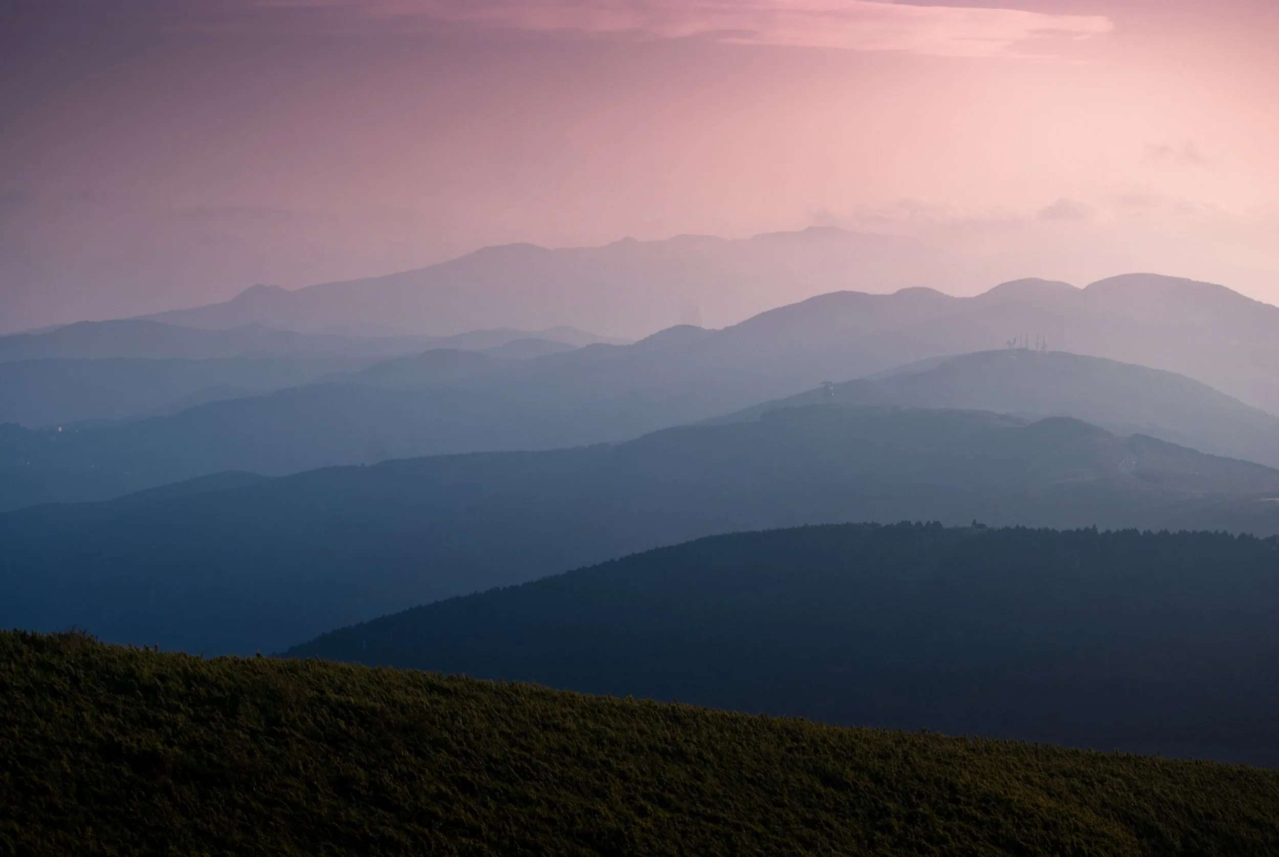 Multiple layers of mountain ranges fading into the distance during sunset, with a soft pinkish-purple sky and a green hill in the foreground.