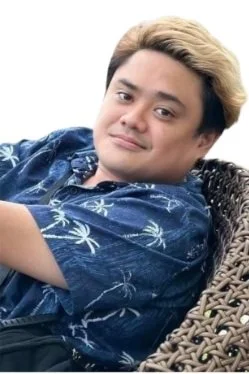 Young man with styled blonde hair wearing a blue Hawaiian shirt, sitting on a wicker chair, smiling at the camera.