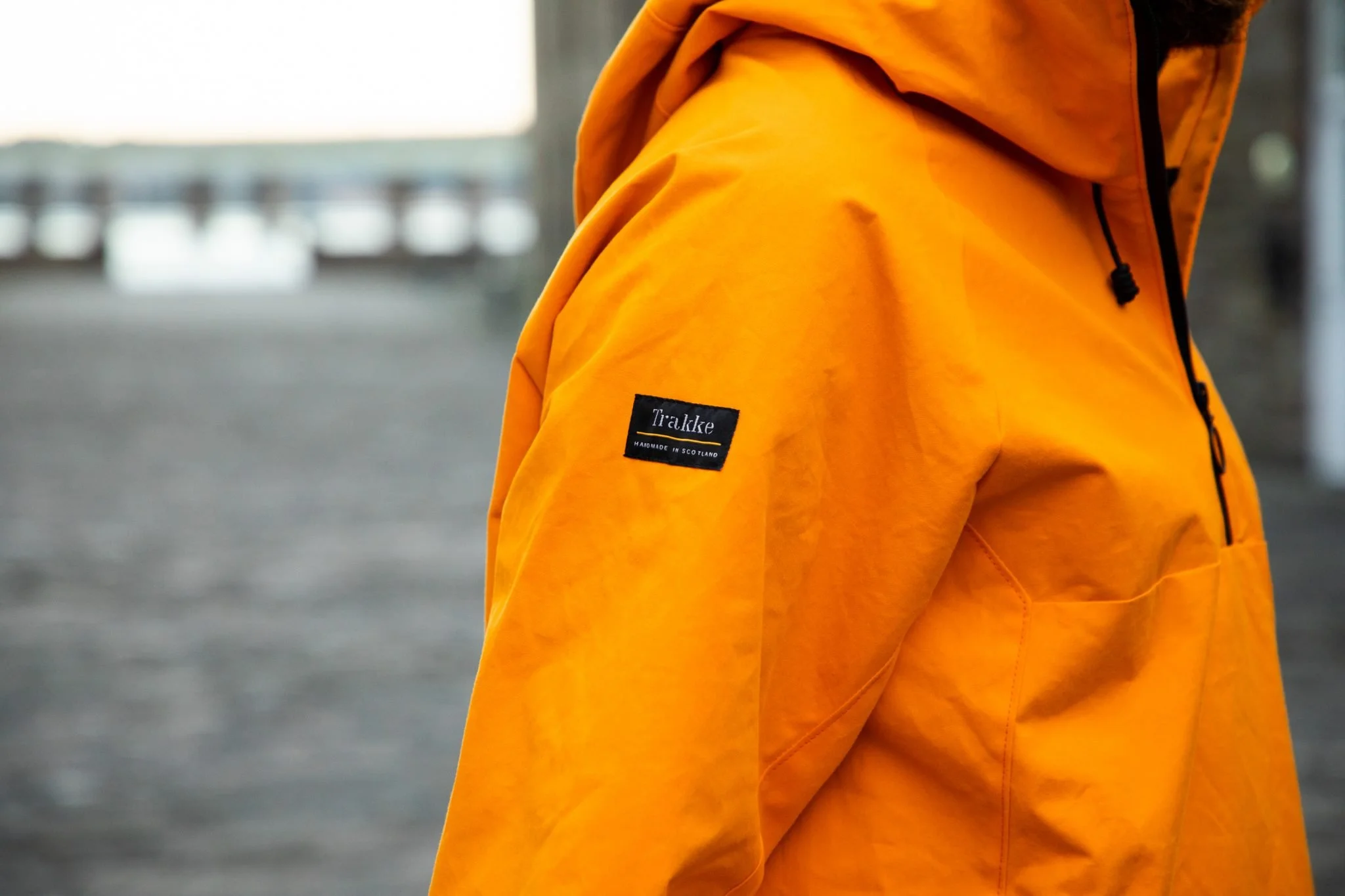 A person wearing a bright orange jacket, with a black label reading "Trakke Handmade in Scotland" on the sleeve, standing outdoors with a blurred background.
