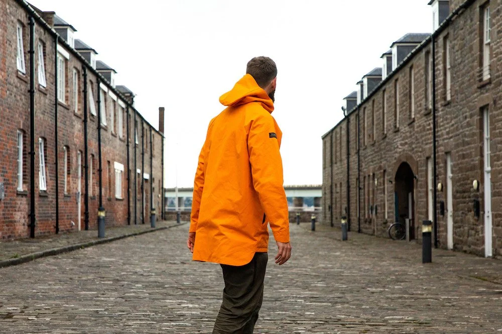 A man wearing an orange jacket walking down a cobblestone street between brick buildings.