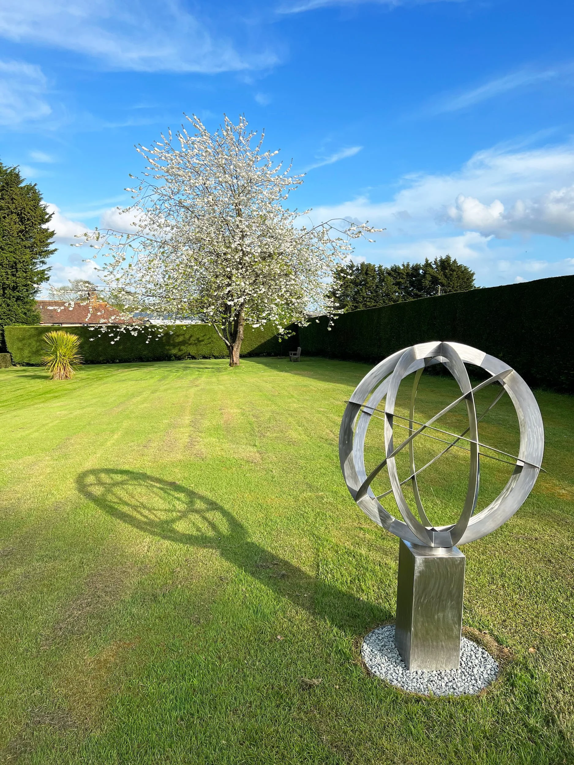 A bespoke contemporary stainless steel armillary sphere sculpture with interlocking metal rings sits atop a matching rectangular plinth on gravel in a sunlit green lawn framed by a tall hedge.