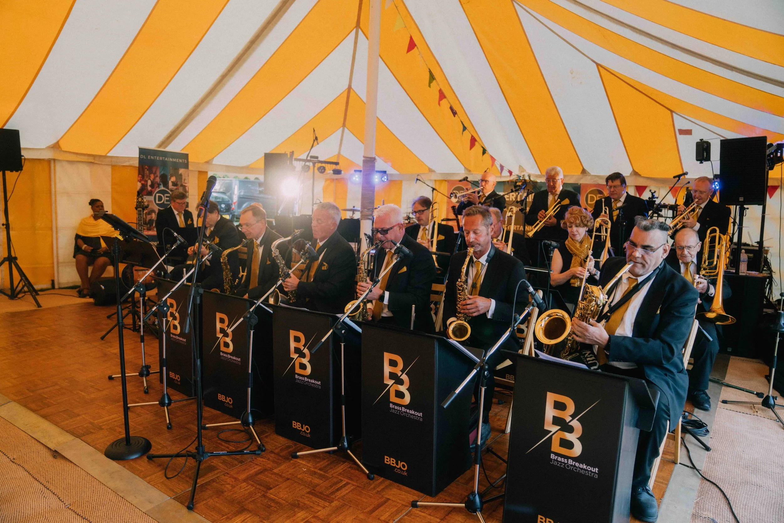 Big band jazz orchestra performing inside a yellow and white striped tent, with musicians in black suits playing saxophones, trumpets, trombones, and other instruments, standing behind black music stands with the logo "Brass Breakout Jazz Orchestra".