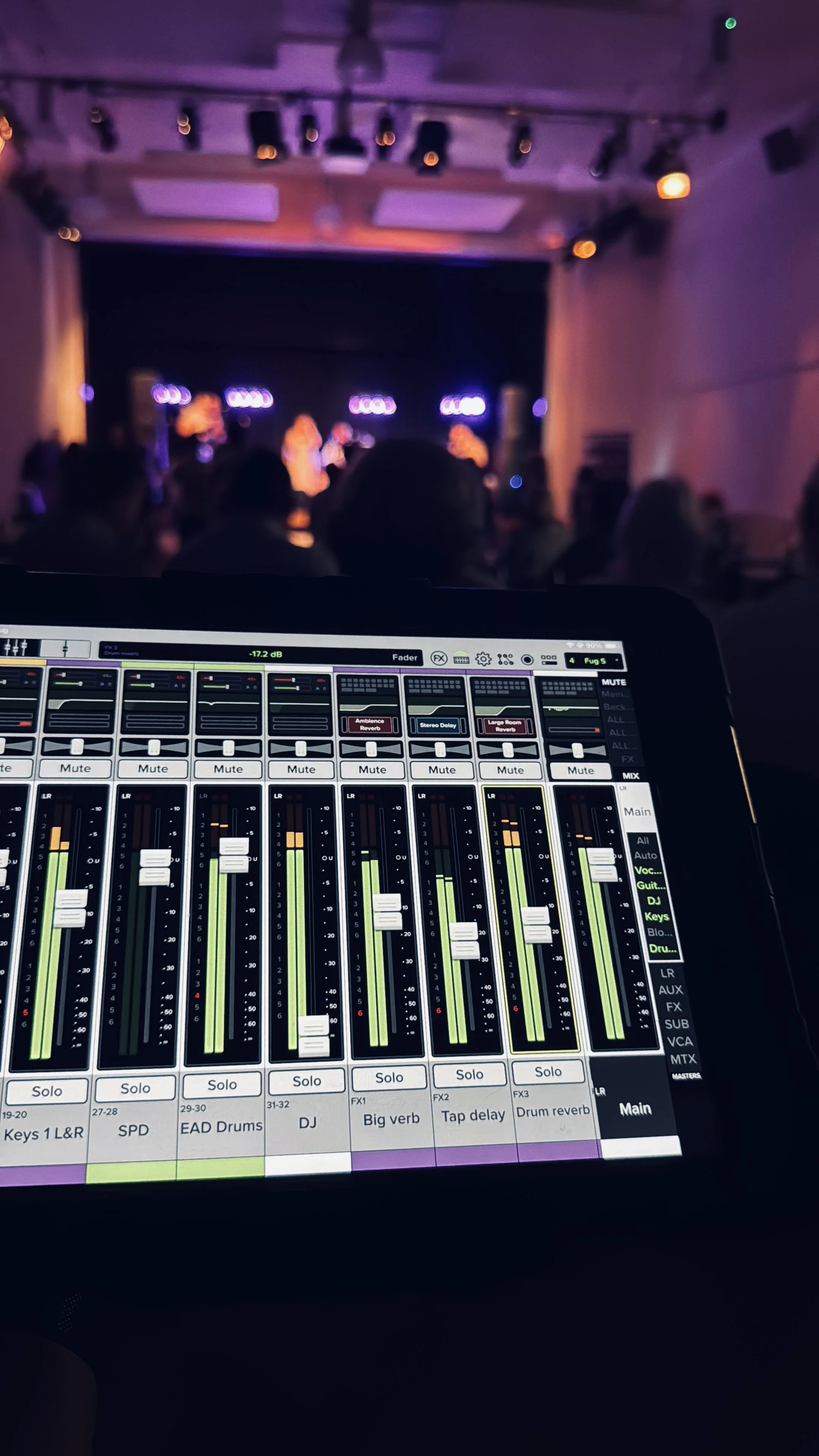 A digital sound mixing console in the foreground with a blurred view of a stage and performers in the background.
