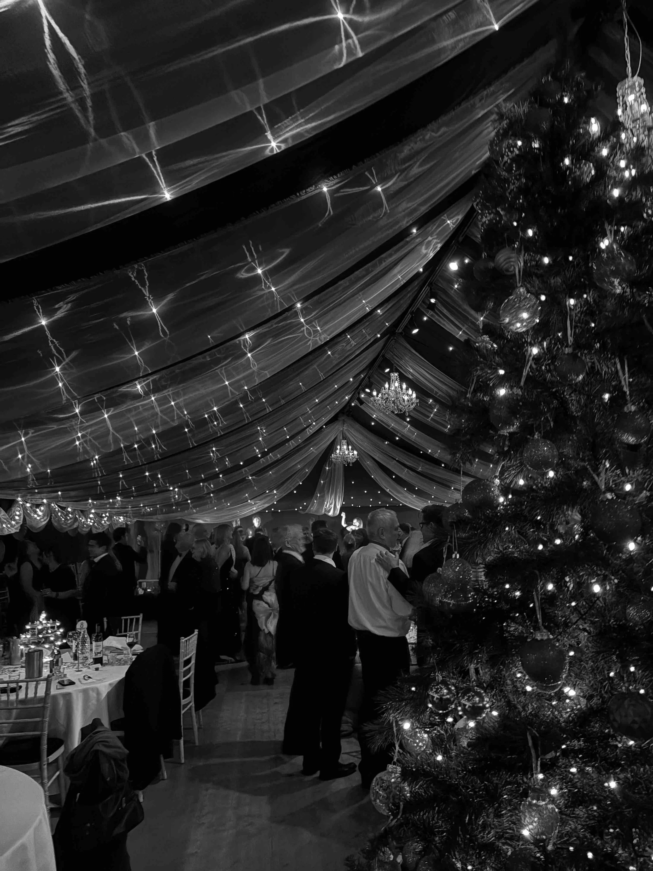 A black and white photo of a large indoor holiday party under a decorated tent. There is a tall Christmas tree with ornaments and lights on the right. People are mingling and dancing in the background. The ceiling has draped fabric and string lights,