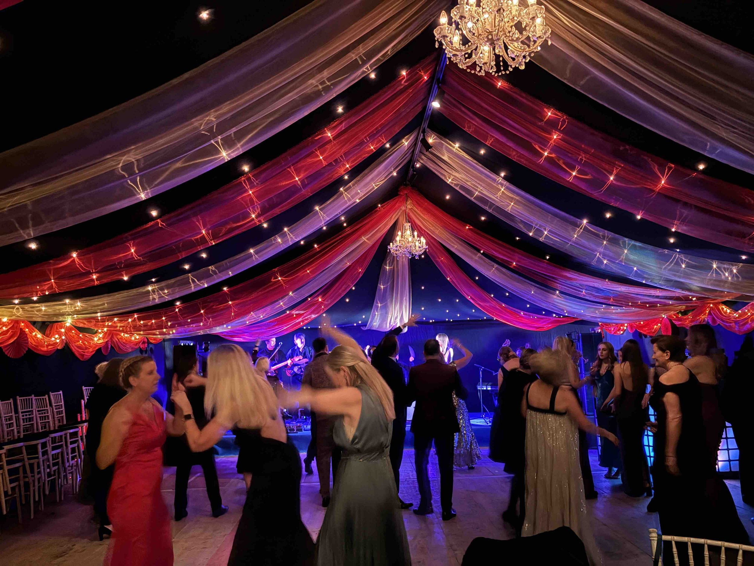 People dancing at a celebration under a decorated tent with colorful draped fabrics and chandeliers.