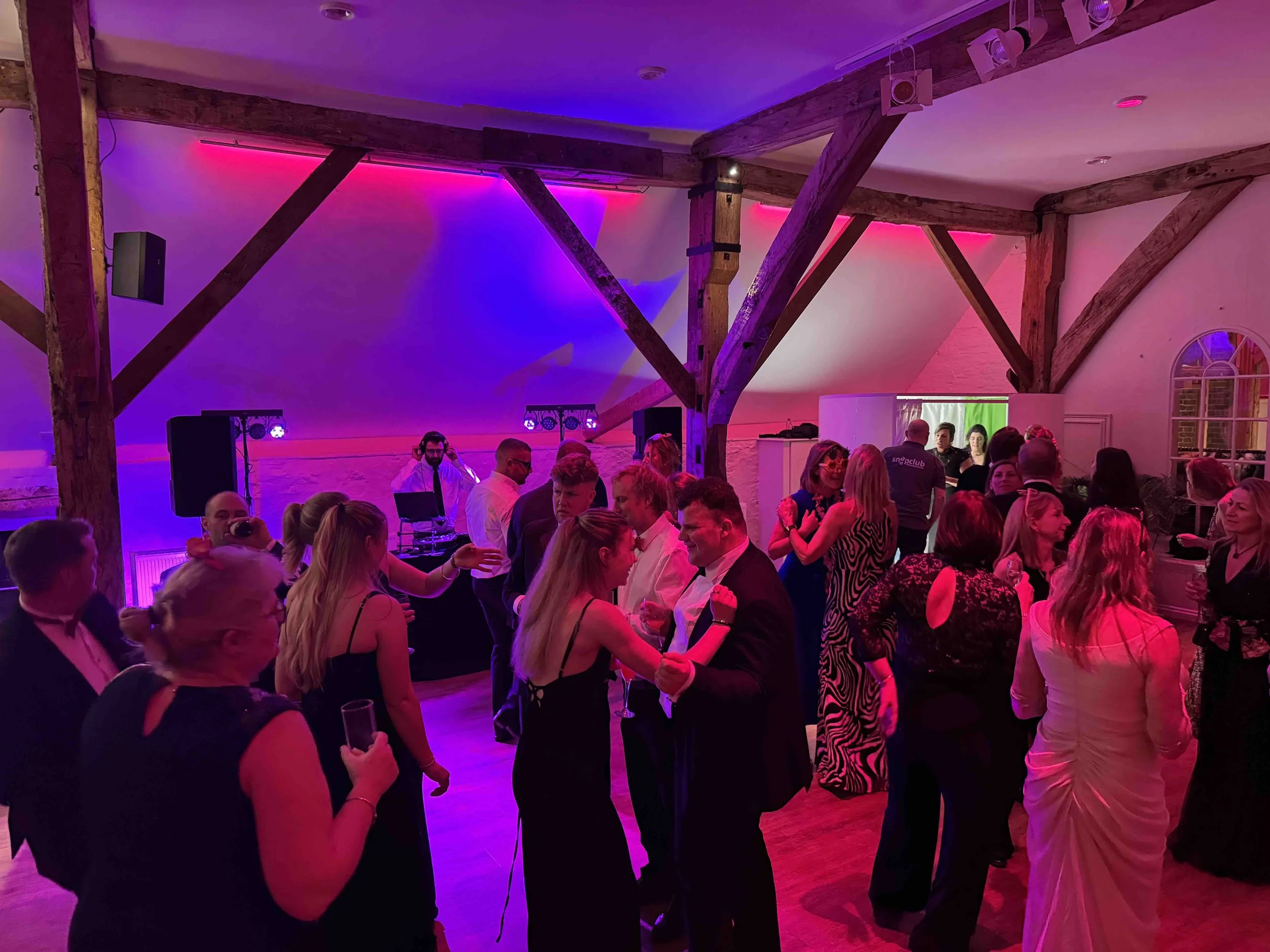 People dancing and socializing at a party or wedding reception in a decorated indoor venue with colorful ambient lighting and a DJ booth.
