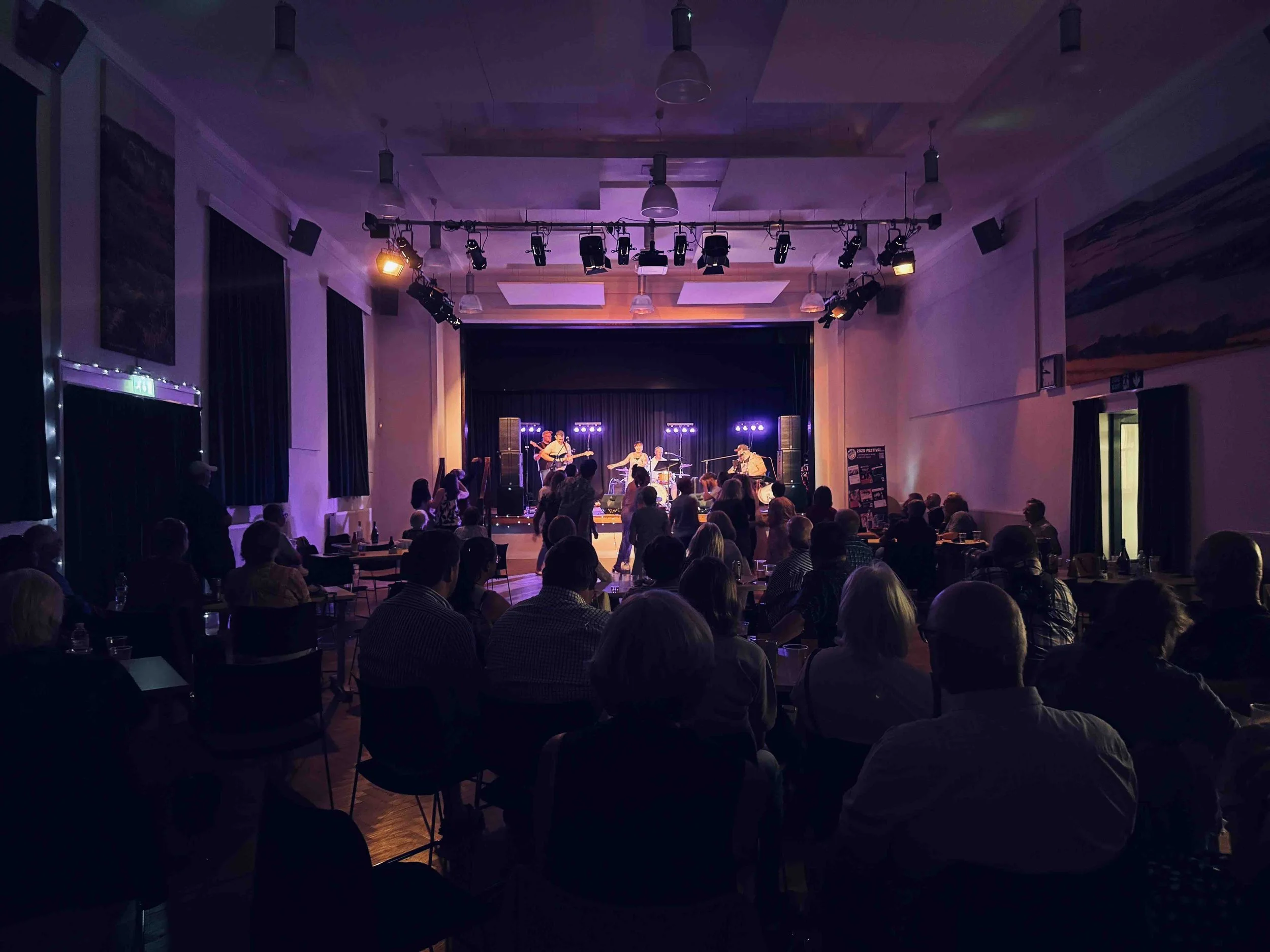 People seated in a darkened concert hall watching a live band perform on stage illuminated with purple and yellow lights.