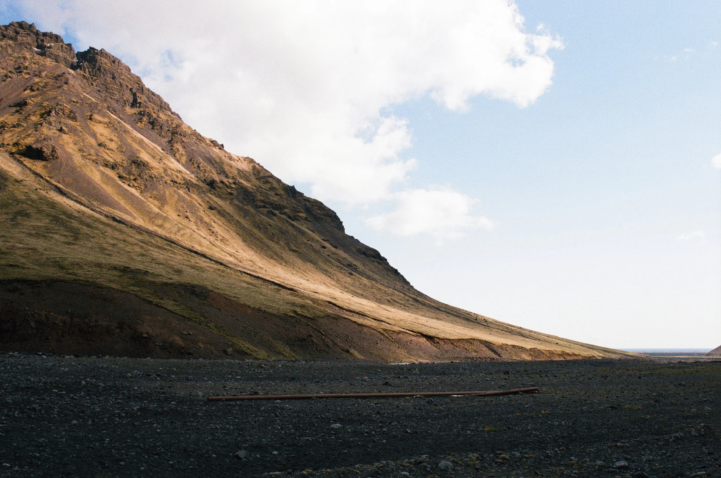 Steep rocky mountain slope rising from a dark volcanic landscape beneath a cloudy sky