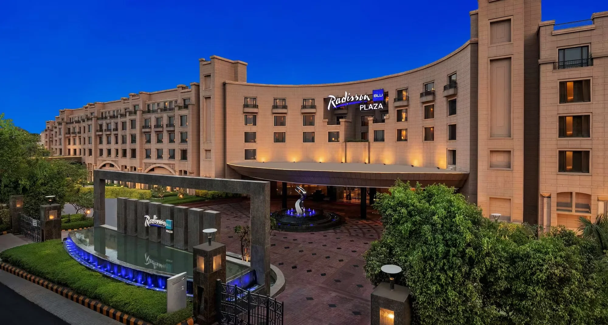 Exterior view of Radisson Blu Plaza hotel at dusk with illuminated sign, fountain, and landscaped greenery.