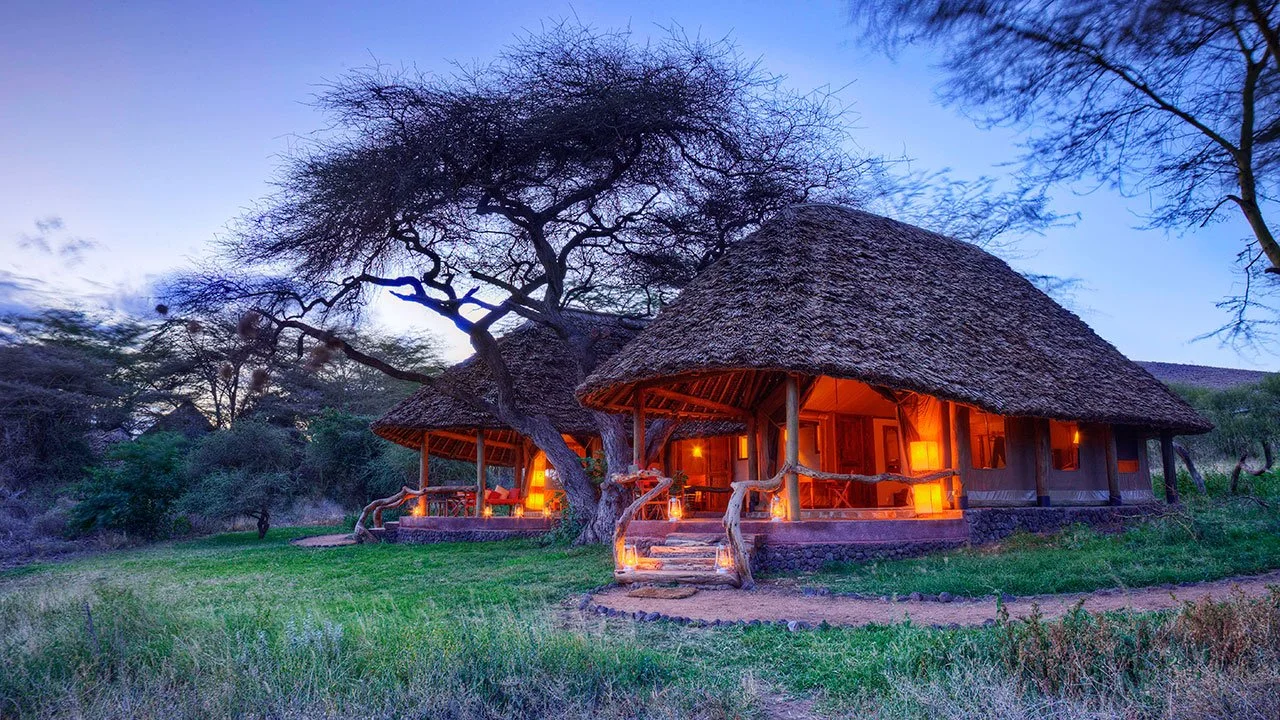 A traditional thatched-roof hut with a porch, illuminated by warm orange lights, surrounded by trees and grass at dusk in a savanna landscape.