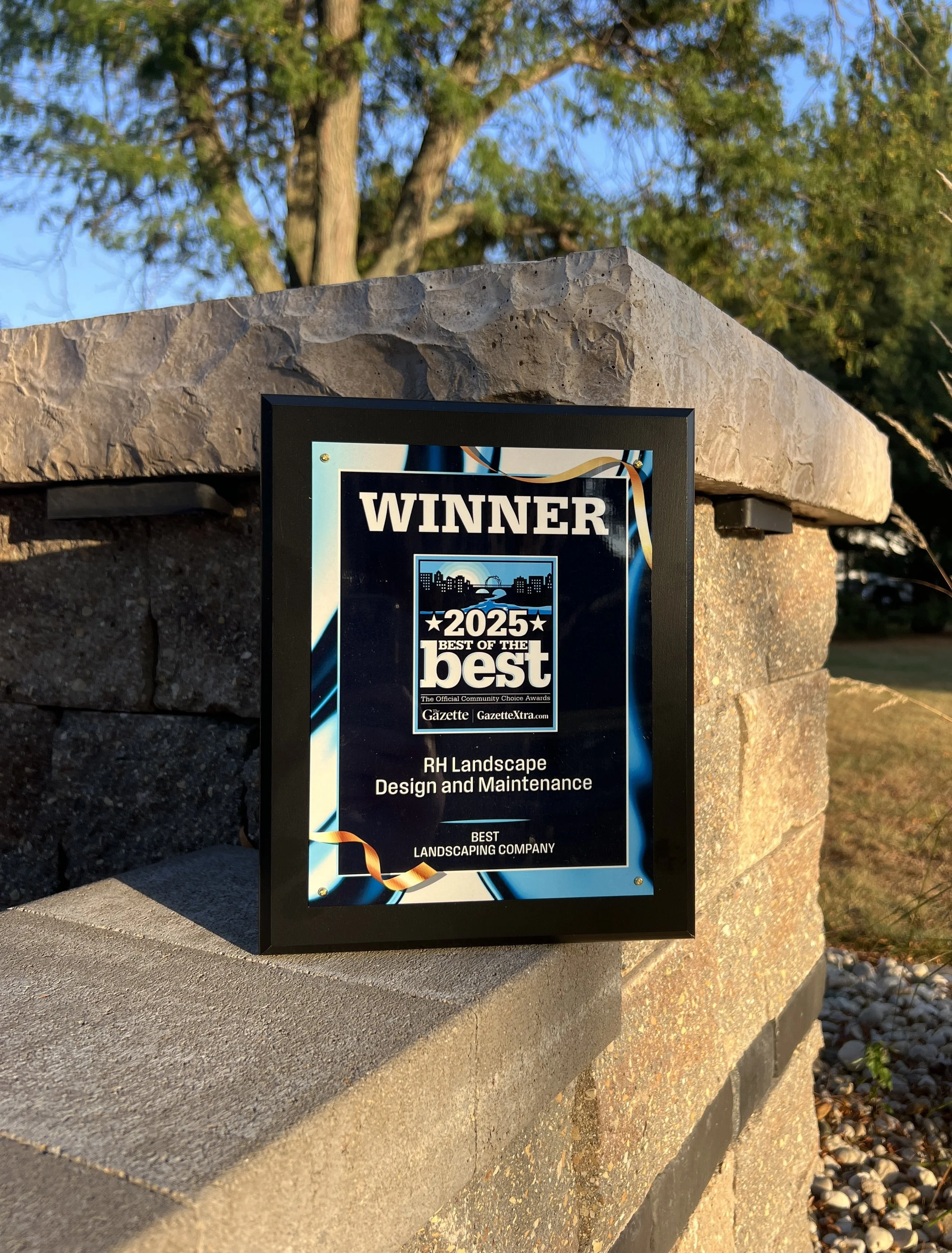 A framed award plaque placed on a stone wall, indicating that RH Landscape Design and Maintenance is the winner of the 2025 Best of the Best Community Choice Award for Best Landscaping Company.