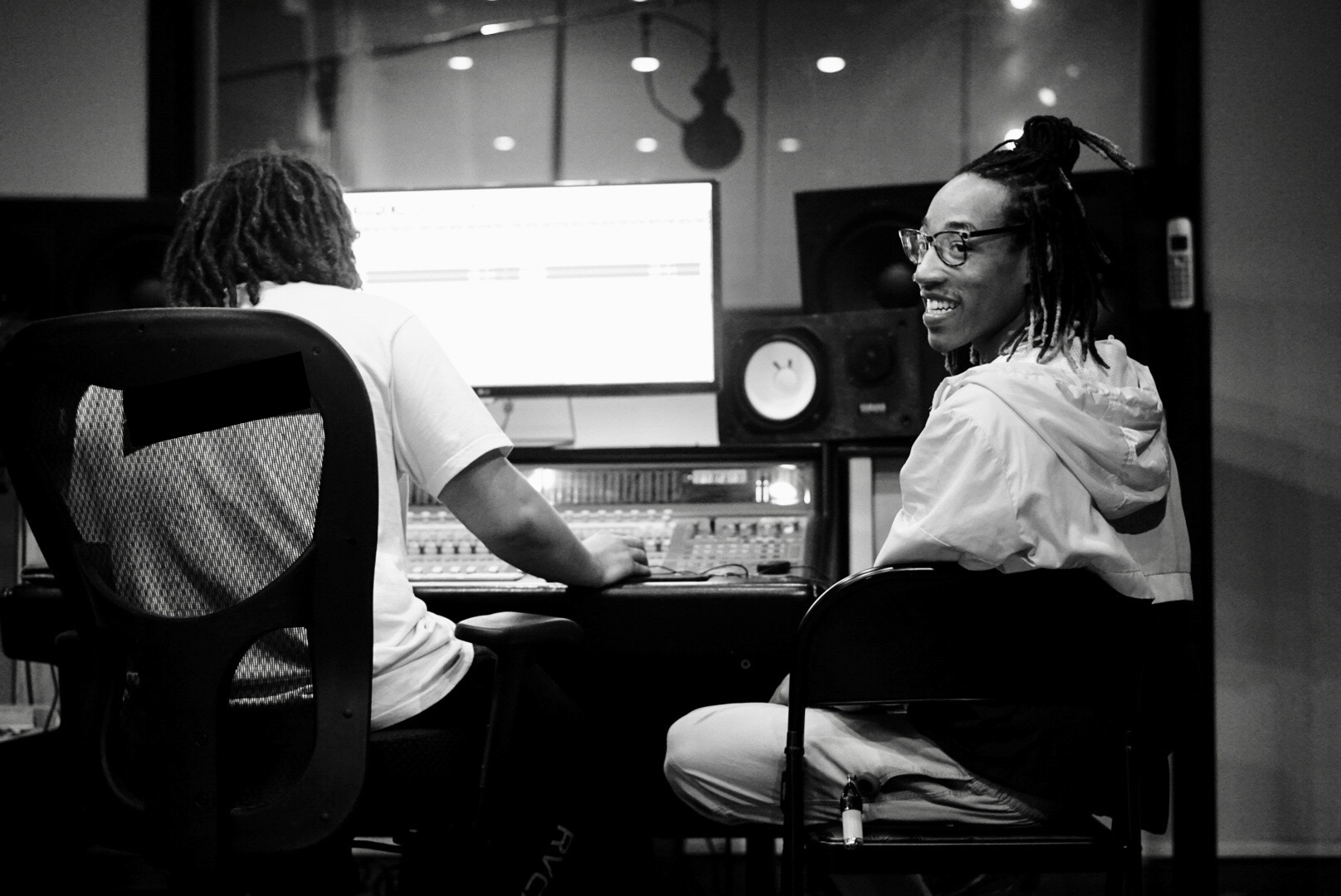 Two people sitting in a music studio, one facing computer screen, audio equipment visible.