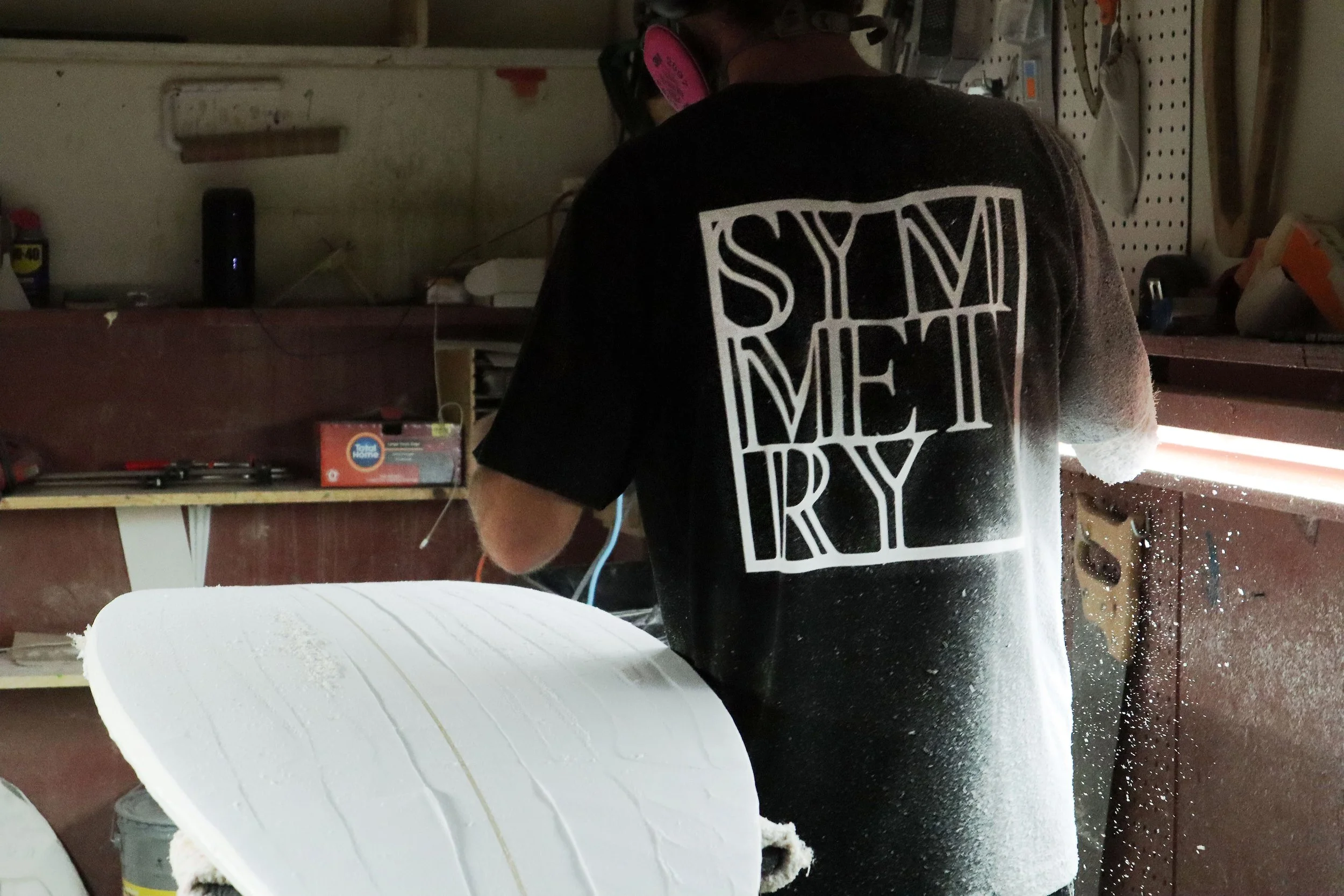 Person shaping a surfboard in a woodworking workshop, wearing a black t-shirt with white text. Dust particles in the air and tools on shelves are visible.