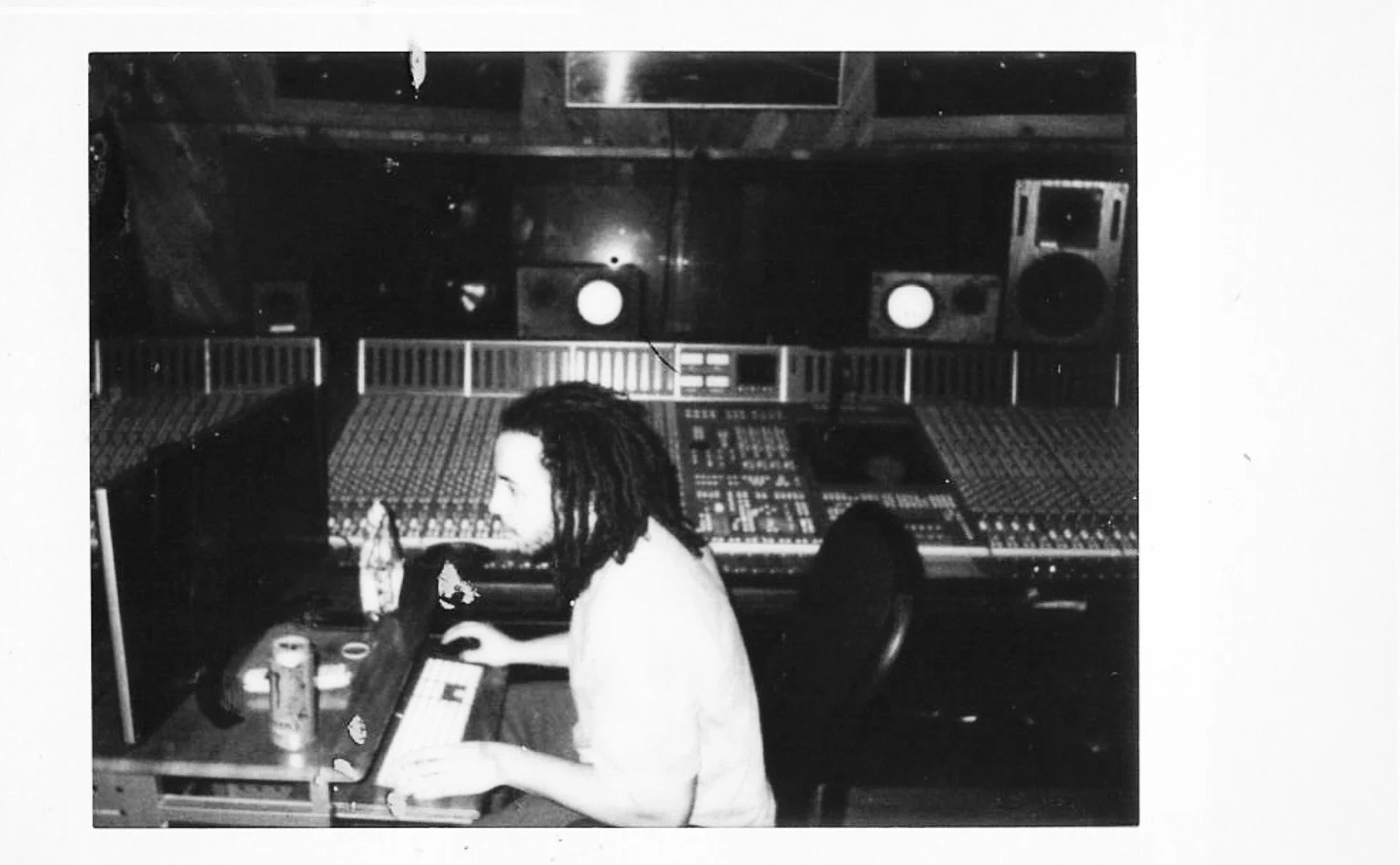 In a recording studio, a man with dreadlocks works on a computer keyboard at a desk with a beverage can and a water bottle. Behind him is a large mixing console, with speakers and acoustic panels on the walls.