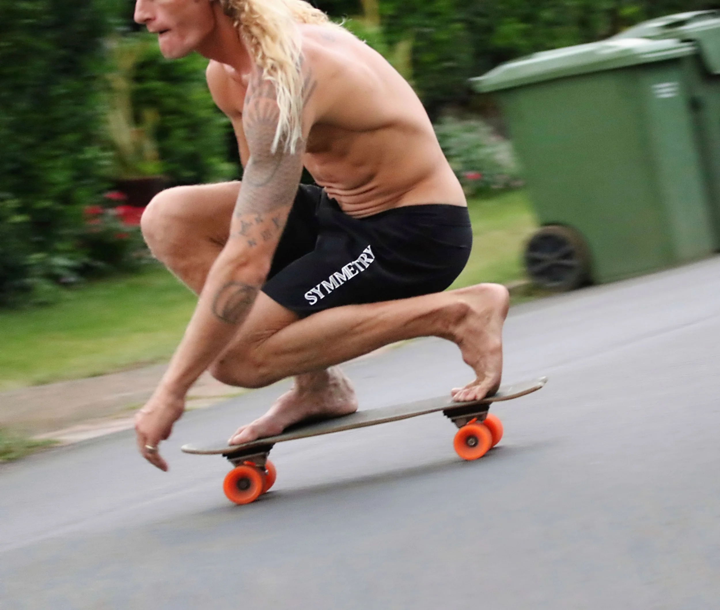 A shirtless man with tattoos crouching on a skateboard, speeding down a suburban street with a green trash bin in the background.
