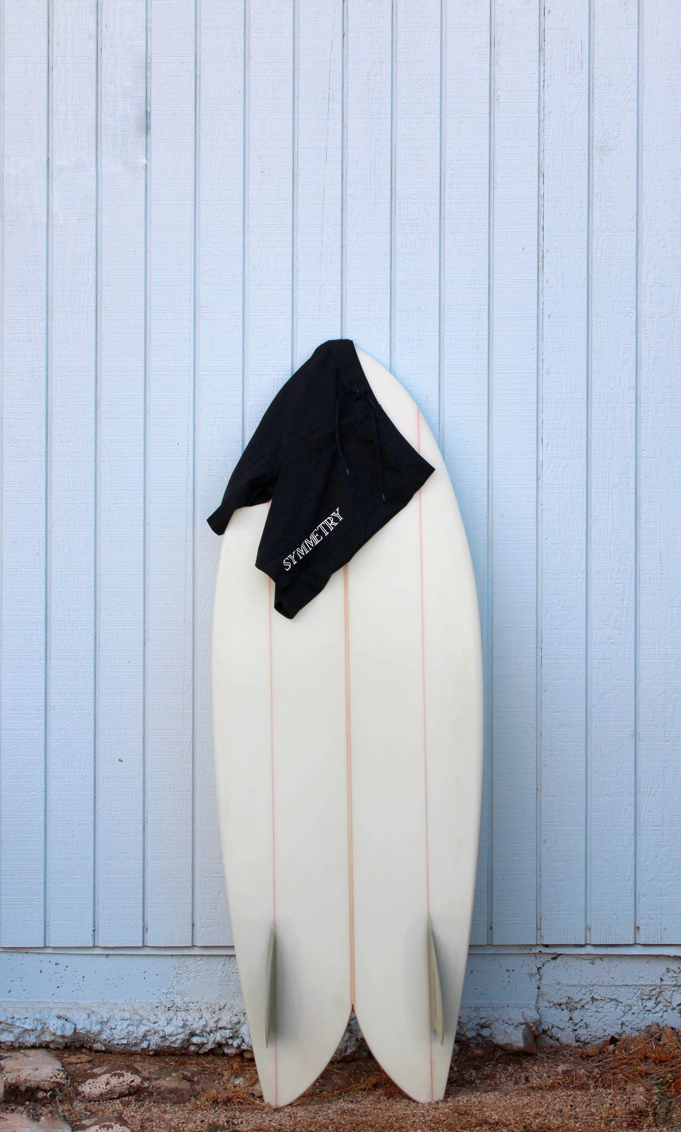 Surfboard and black shorts that read 'SYMMETRY' leaning against light blue wall.