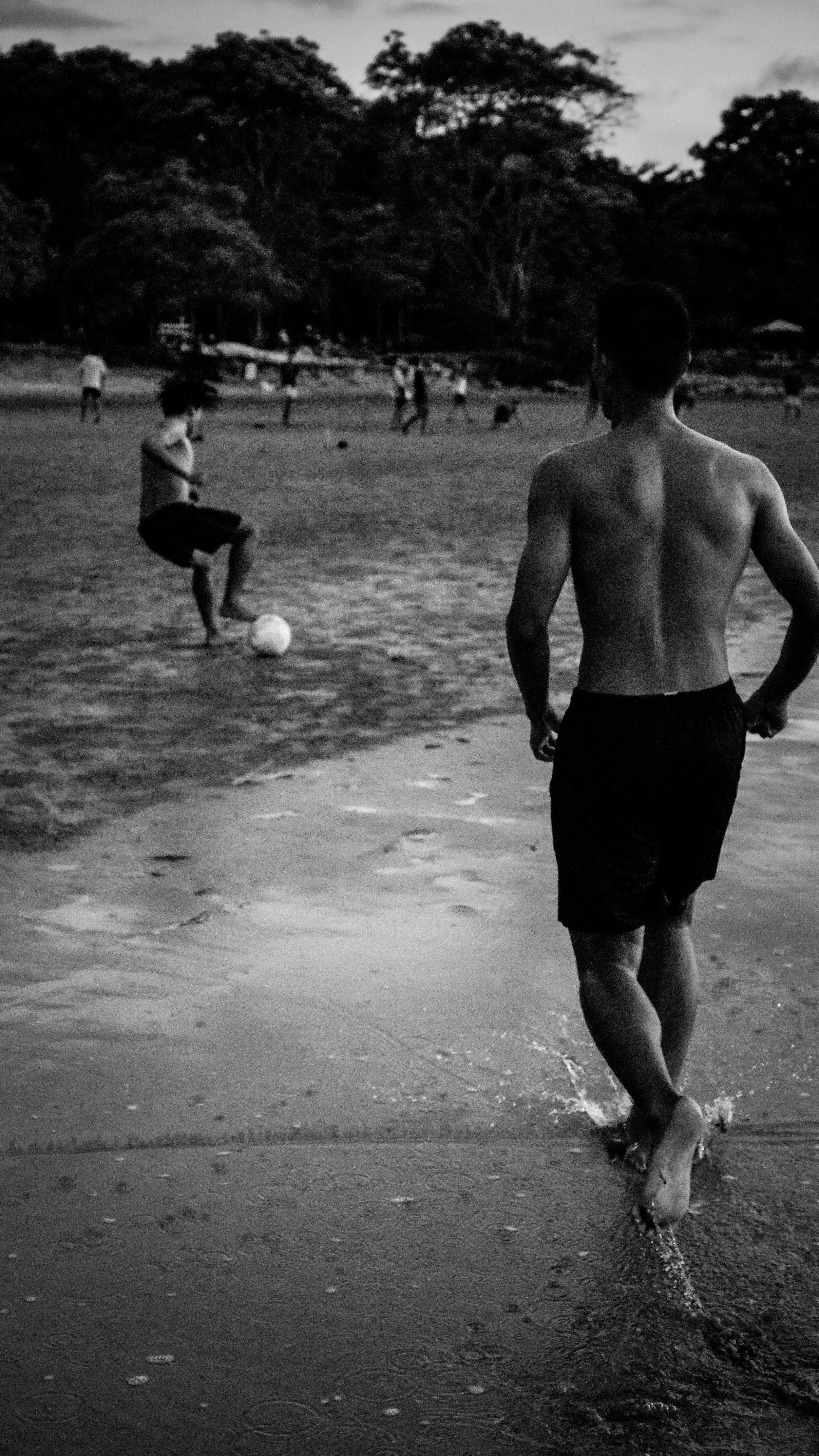 A young man runs into the water at the beach, with children playing soccer nearby and trees in the background, captured in black and white.