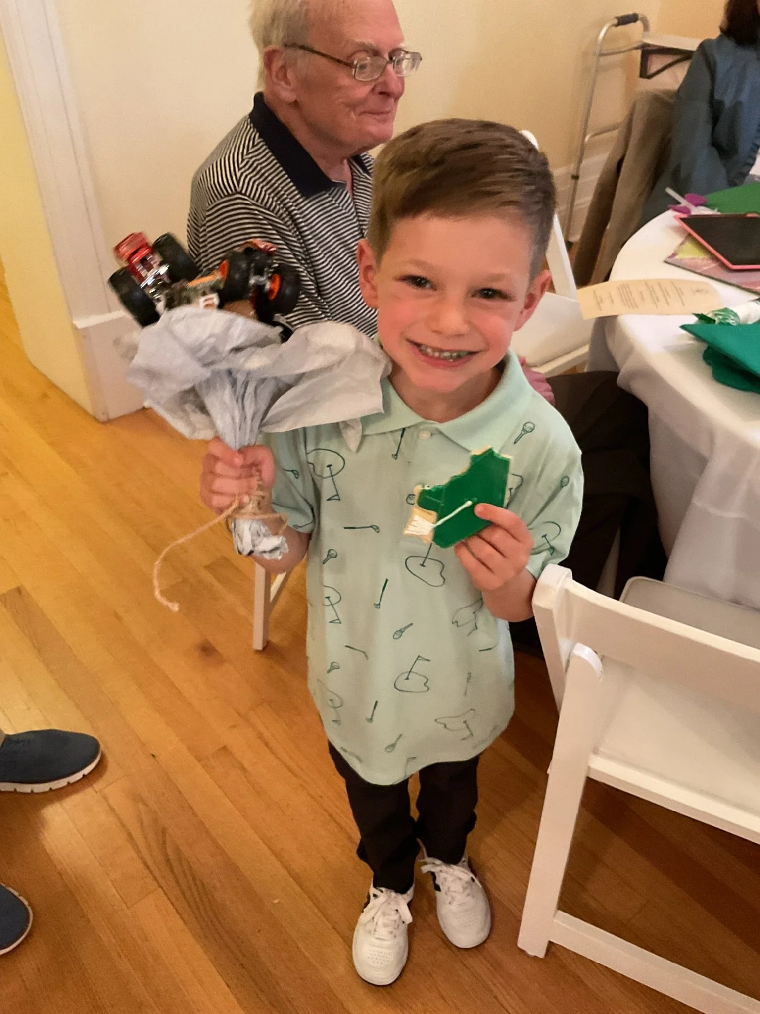 A young boy holding a toy car wrapped in tissue paper, smiling at the camera, standing on a wooden floor with an older man sitting at a table behind him.