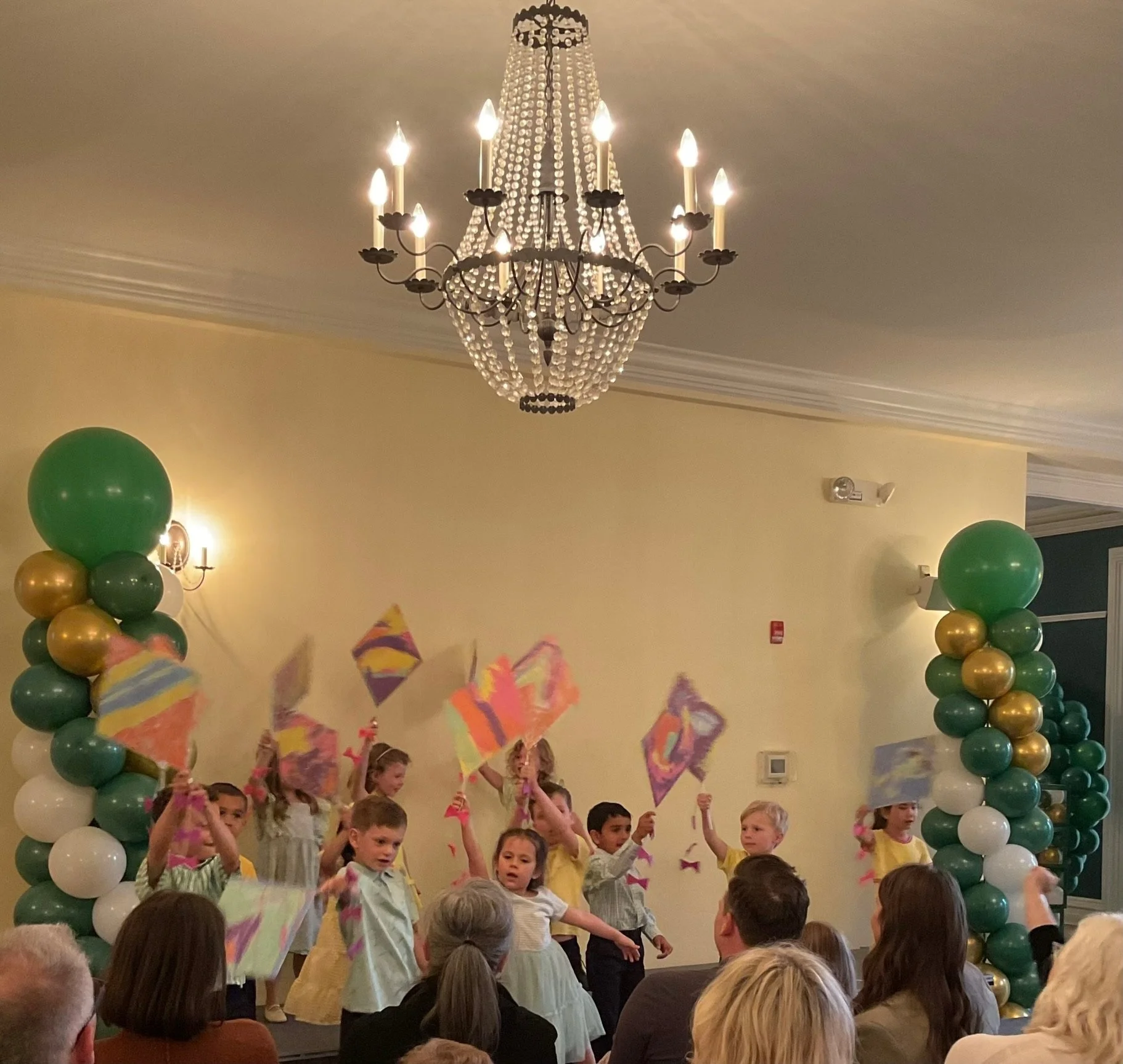 Children performing or participating in a celebration or event with colorful paper flags, amid large balloon columns in green, white, and gold, with a chandelier hanging from the ceiling in a decorated room.