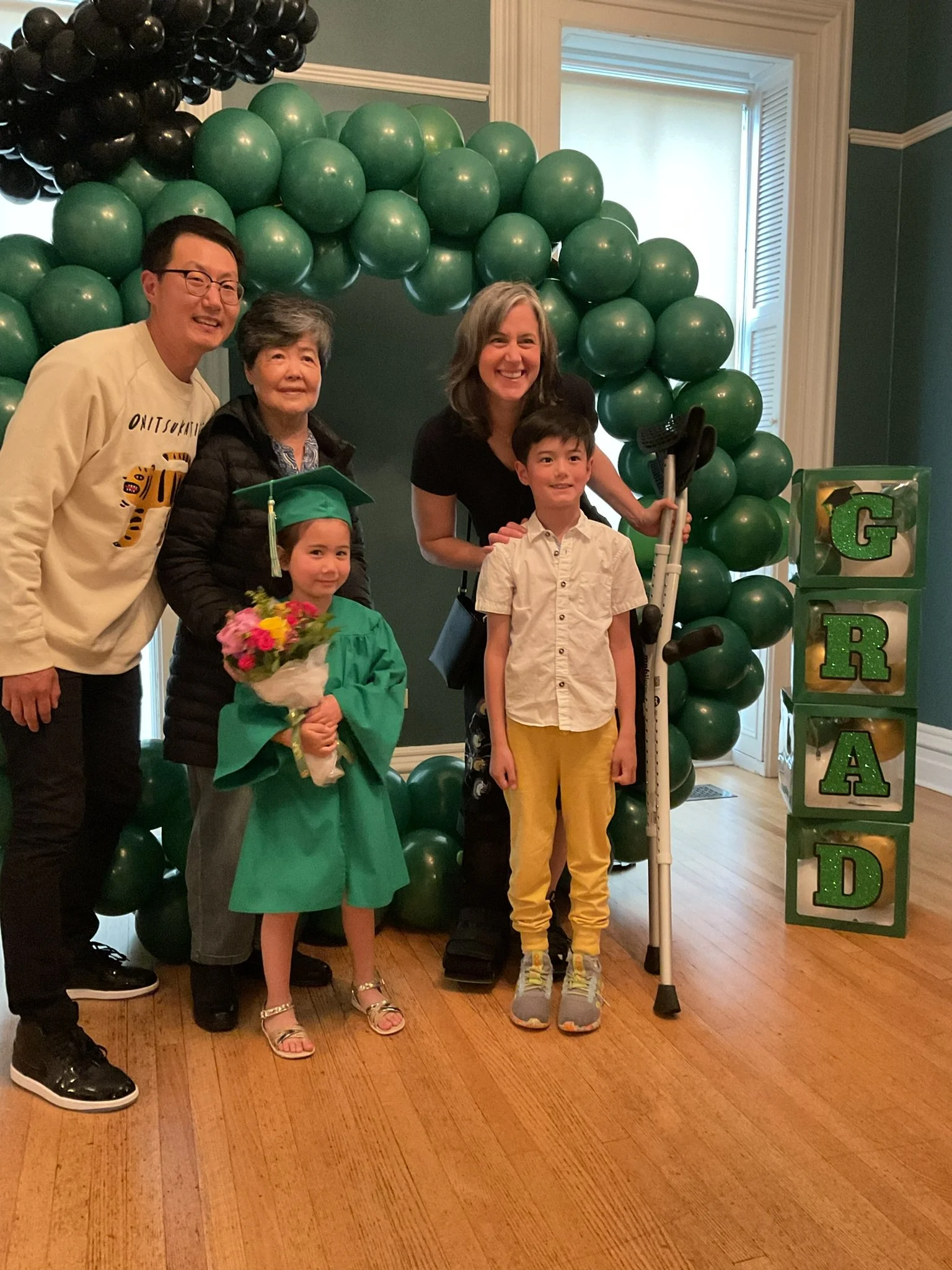 Group of five people, including a young girl in a graduation cap and gown holding flowers, a woman and three men, standing in front of a balloon arch and a decorated frame that spells out "GRAD." They are indoors with wooden floor and a window in the