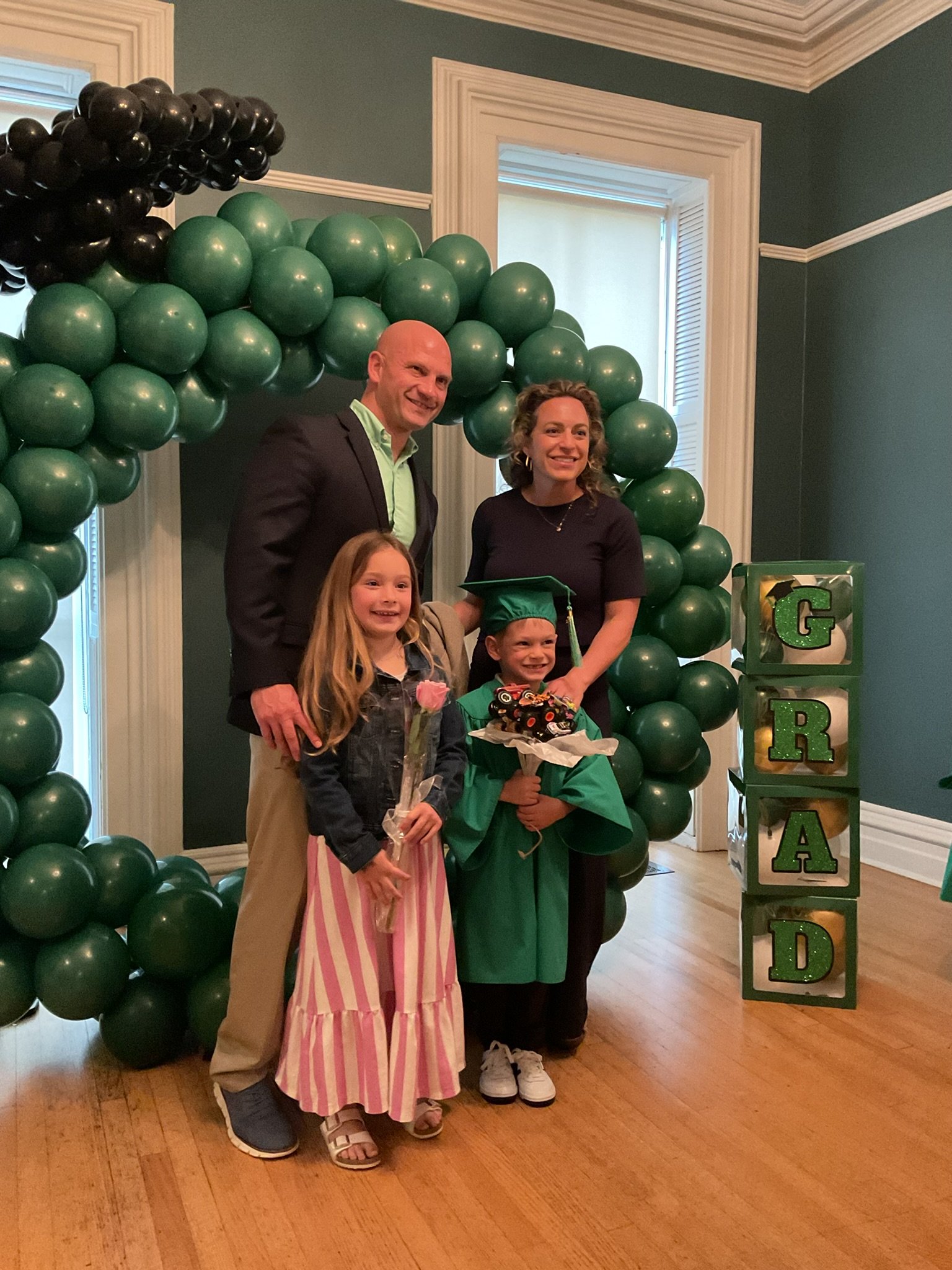 A family celebrating a graduation indoors, standing in front of a balloon arch with green and black balloons. The graduate, wearing a green cap and gown, is holding a bouquet and a decorated cake. Two other children and two adults are smiling and pos