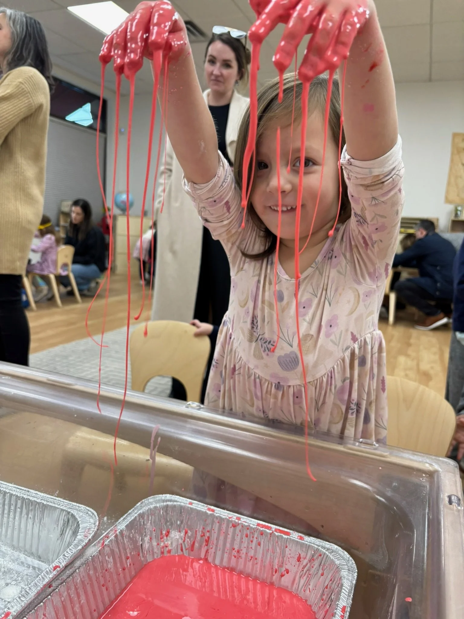A young girl is playing with red slime, lifting it with her hands and stretching it over her face, in a room with people in the background.