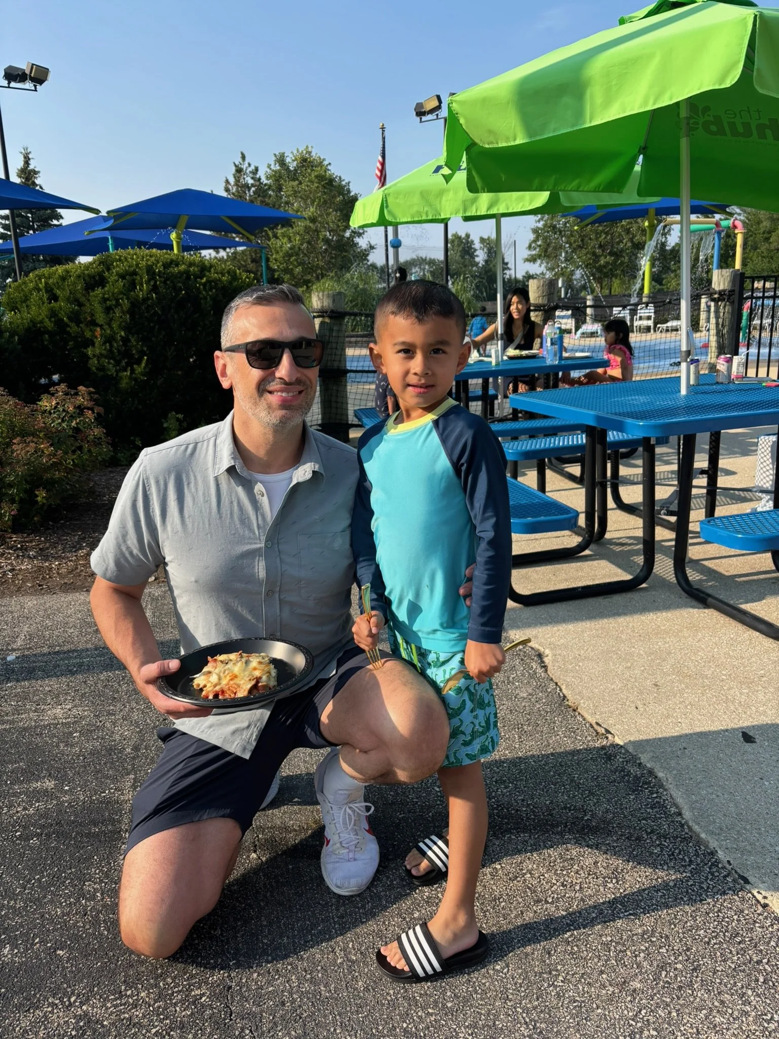 A man kneeling on one knee next to a young boy at an outdoor pool area. 