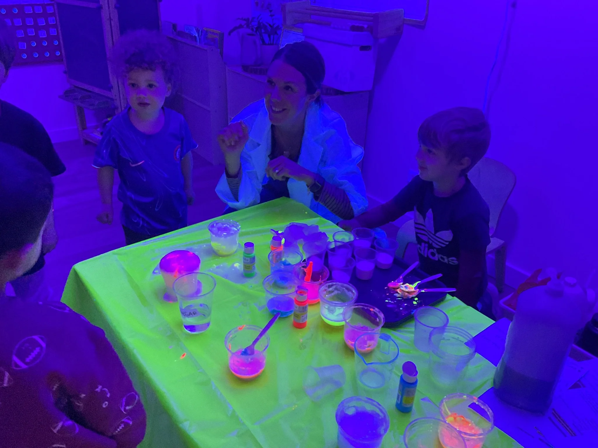 Children and an adult are around a table with glow-in-the-dark slime and cups, in a room with ultraviolet lighting