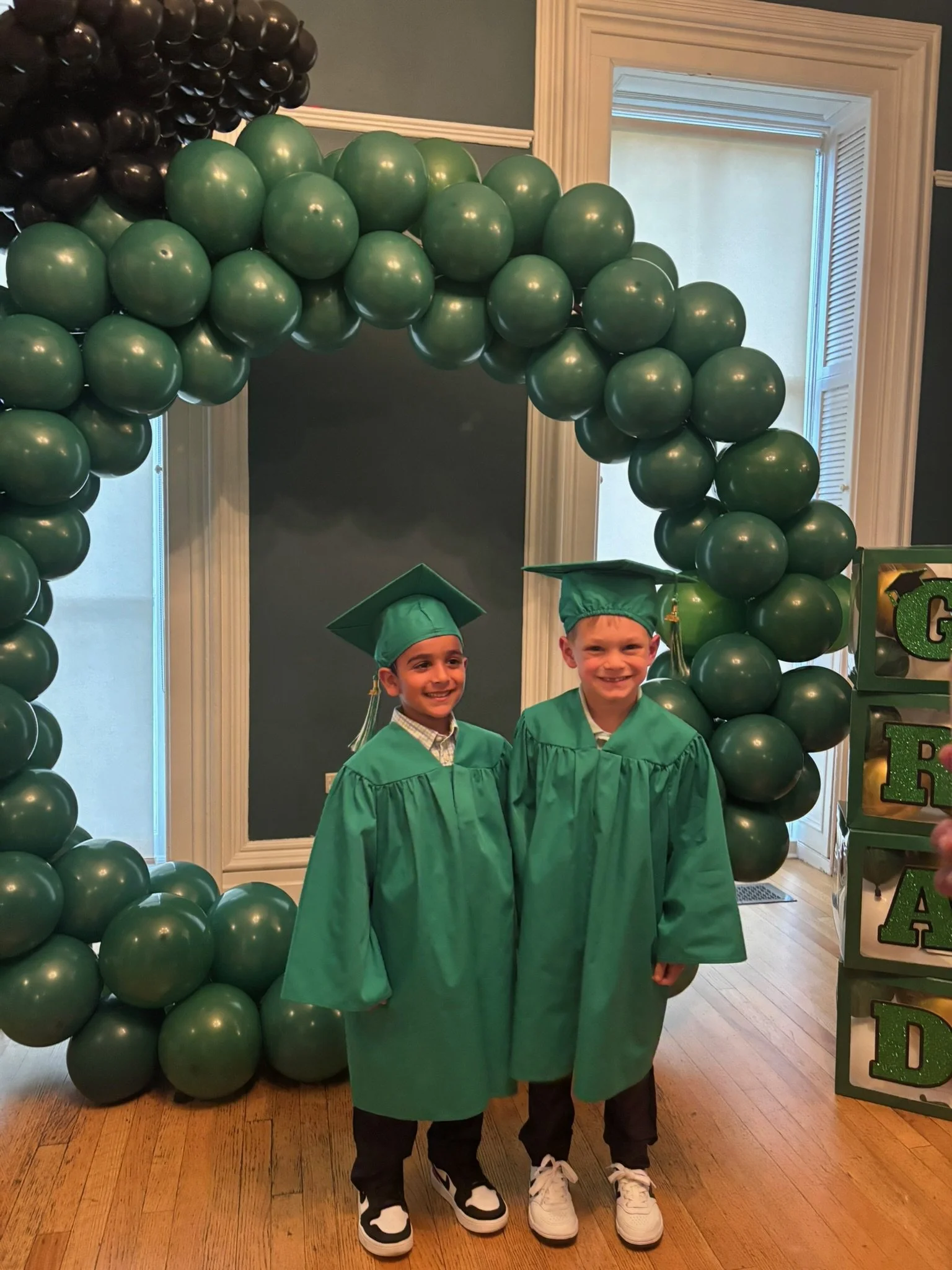 Two young boys wearing green graduation caps and gowns standing in front of a balloon arch at a graduation celebration indoors with wooden floors and a decorative window frame.