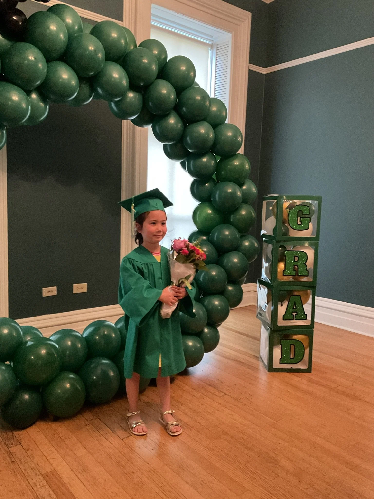 A young girl wearing a green graduation cap and gown holding a bouquet of flowers, standing in front of a graduation arch made of green balloons and a decorative block column spelling "GRAD".
