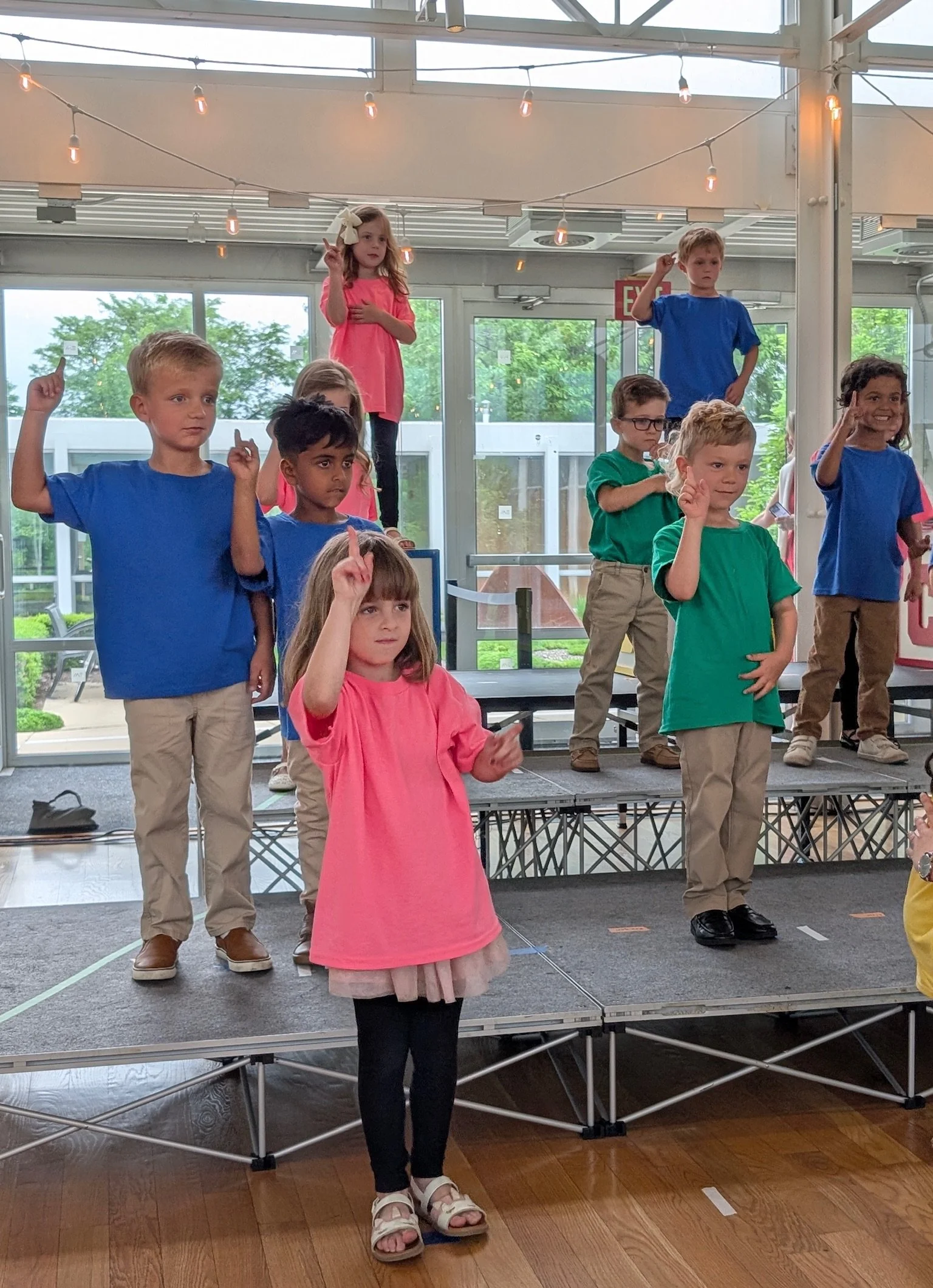 Children standing on a stage, some raising their hands, in a room with large windows and string lights hanging from the ceiling.